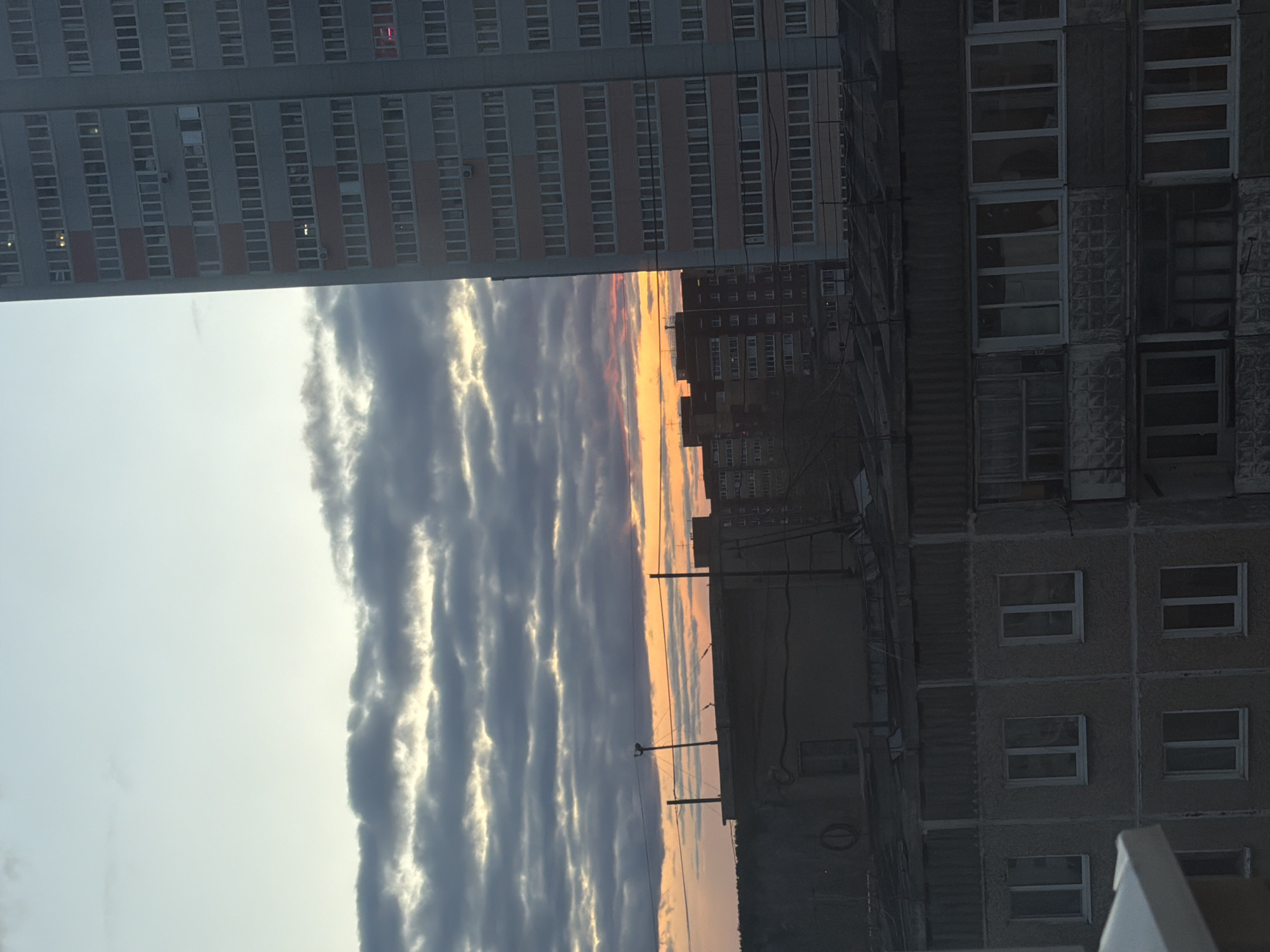 The image depicts a view in the city of Perm, Russia, characterized by a sky with striated clouds at sunset framed by the architecture of apartment buildings. On the left, a multi-story apartment building is visible with rows of windows. On the right, a different apartment building is seen with a blocky facade and windows. The sky is the central focus, with a gradient of colors transitioning from light blue to orange and pink at the horizon, indicating sunset. The striated clouds are positioned vertically. Utility poles with wires are also visible in the scene.