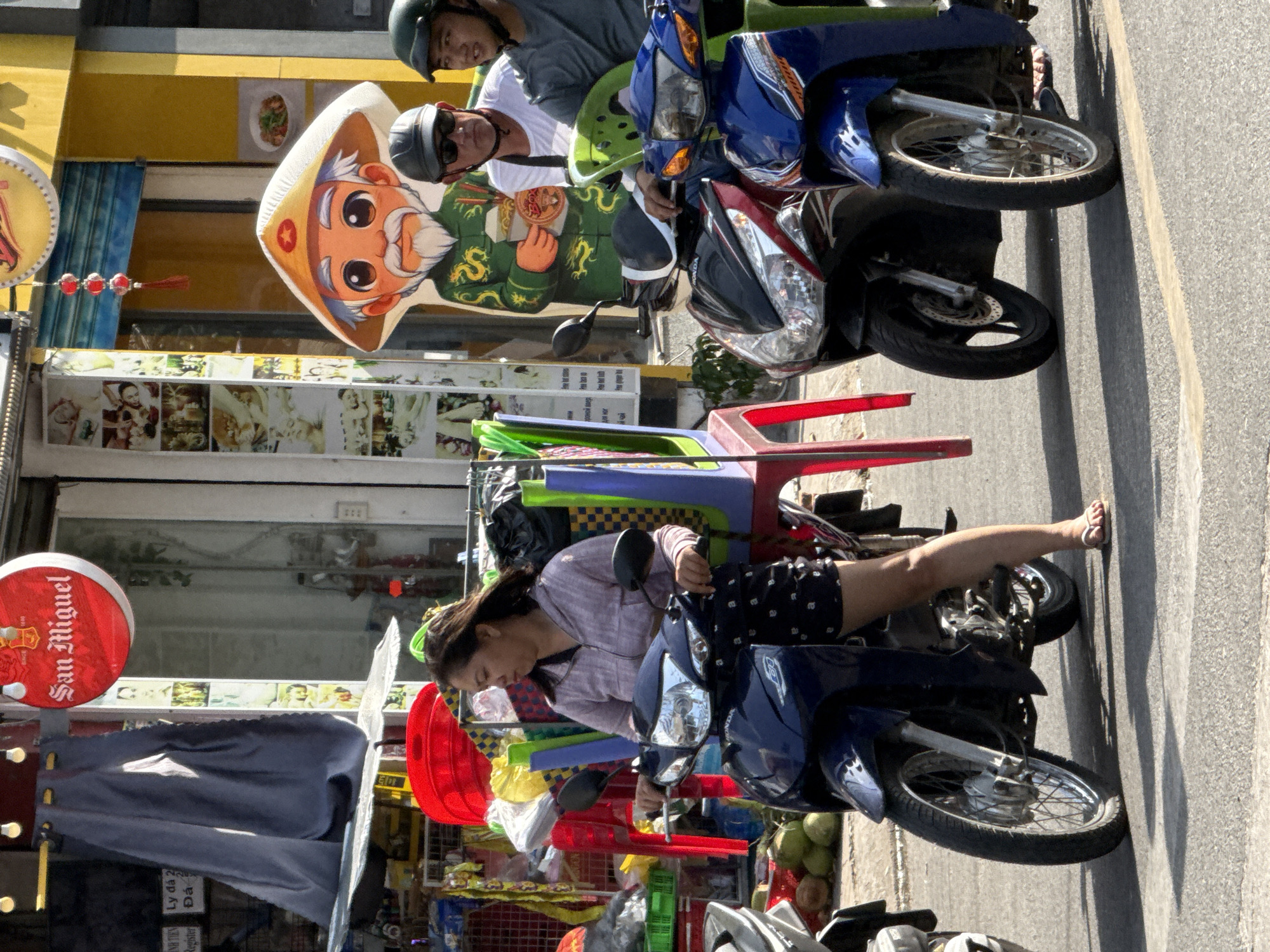 In Bac Nha Trang Ward, Vietnam, a woman rides a dark blue Honda motorcycle. She is wearing a light purple hooded jacket and black shorts with a white polka dot pattern. Her hair is dark brown and blown to the side by the wind. In front of her on the motorcycle is a bright red plastic chair and what appears to be a green plastic seat, both loaded with items. To her left, another motorcycle is partially visible.
Behind her, a large, cartoonish cutout of an elderly man with a conical hat stands. He is wearing a green robe with gold dragons and holding a bowl of noodles. To the right of the cutout, two men are seated on a motorcycle. The man in the foreground is wearing a gray helmet and sunglasses, with a white t-shirt. The man behind him is wearing a dark helmet and a gray t-shirt.
The scene takes place on a paved street in front of a building with a yellow facade. A red circular sign with "San Miguel" in white lettering is visible above the entrance. A string of red lanterns hangs from the building. Various signs with text and images are displayed on the wall of the building. In the background, there are shadows cast on the ground from the sun. To the left of the woman on the motorcycle, a stack of green coconuts is visible on the ground.