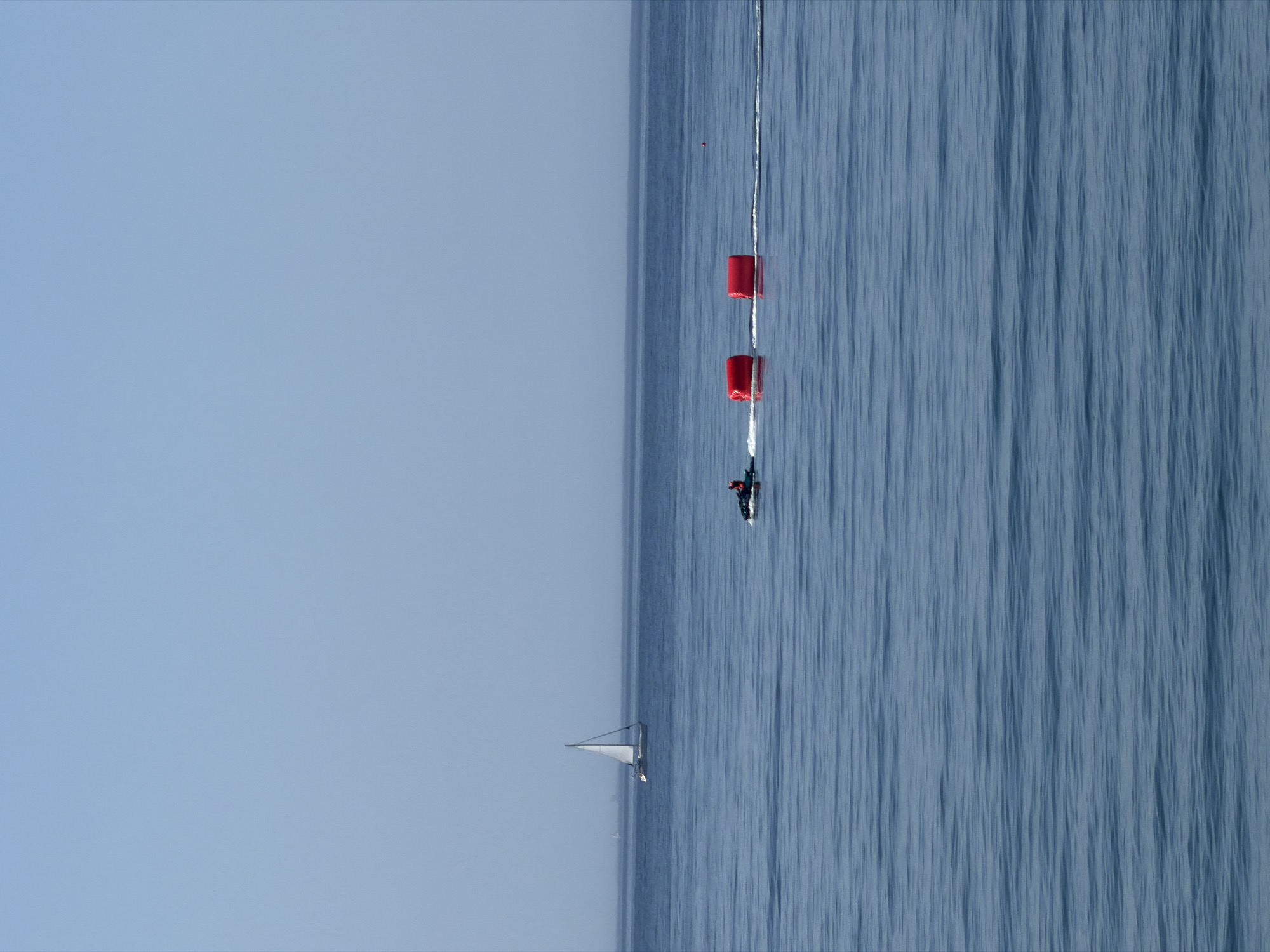 An unobstructed, wide shot captures a clear blue sky meeting the calm blue sea. In the mid-distance, a lone white sailboat glides across the water, its sail full. Closer to the foreground, a person in a red jacket rides a jet ski, leaving a wake behind them. Two large, cylindrical red buoys are positioned in a line to the right of the jet ski, with a small red marker buoy further to the right. The horizon is a thin, hazy line separating the sea from the sky. The scene is likely offshore of Estepona, Spain, given its coastal Mediterranean setting.