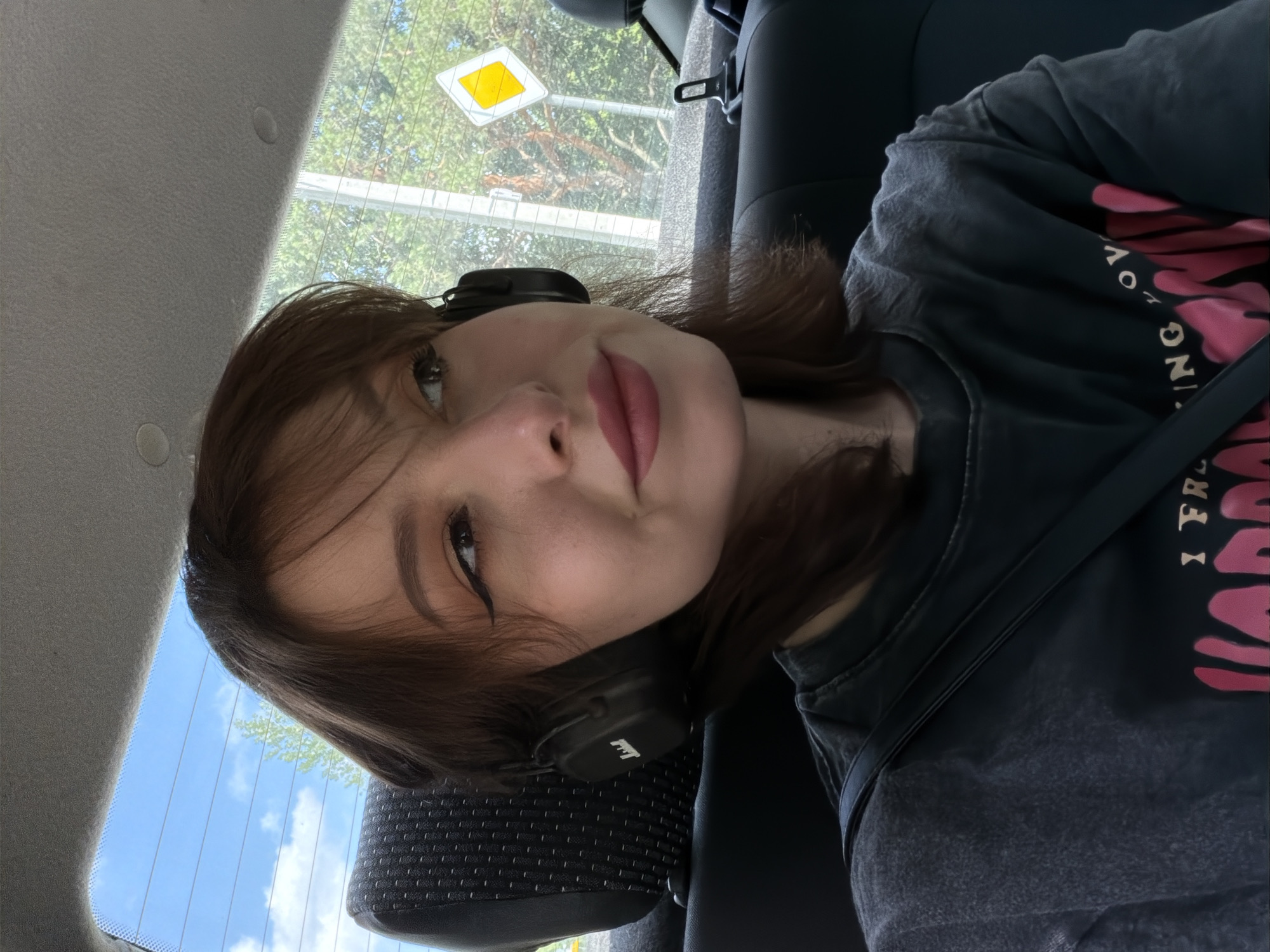 A young woman with brown hair and winged eyeliner is seen from the chest up, seated in the back seat of a car. She is wearing black headphones and a dark, distressed t-shirt with pink lettering. Her lips are painted a matte berry color, and she is looking to her right, with a slight smile. Through the car's rear window, a tree and a yellow diamond-shaped road sign are visible against a backdrop of a blue sky with white clouds. The seat behind her is black with a textured pattern.