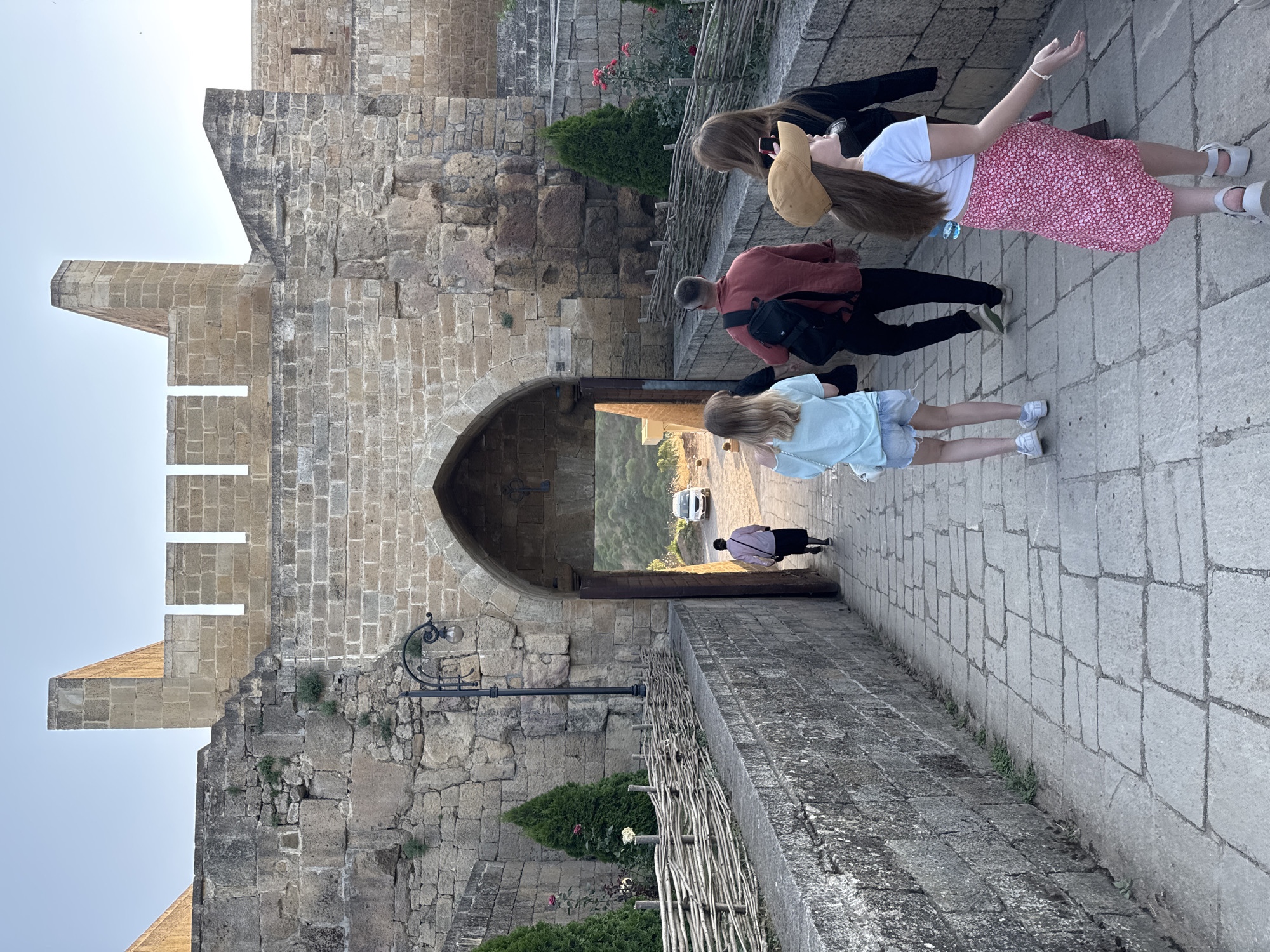 A group of five people are walking across a stone bridge towards an arched gate of an ancient stone fortress. The fortress walls are constructed of large, rough-hewn stones and are topped with crenellations. A lamppost with a single bulb stands on the left side of the bridge. Fencing made of interwoven branches lines the left side of the bridge. Small evergreen trees and some pink roses are planted along the fence.
The people are walking away from the camera, towards the fortress gate. The person in the front right is a young girl wearing a tan baseball cap, a white t-shirt, and a pink floral skirt. Next to her is a person wearing a black top. To their left and further ahead is a man in a red shirt and black pants, with a black backpack. Further ahead, walking through the gate, is a person in a light blue t-shirt and denim shorts. Closest to the gate, walking into it, is a person wearing a pink shirt and a dark skirt. Outside the gate, a white vehicle is visible. The sky above is clear and pale blue.
This image depicts the entrance to the Narikala Fortress in Derbent, Russia.