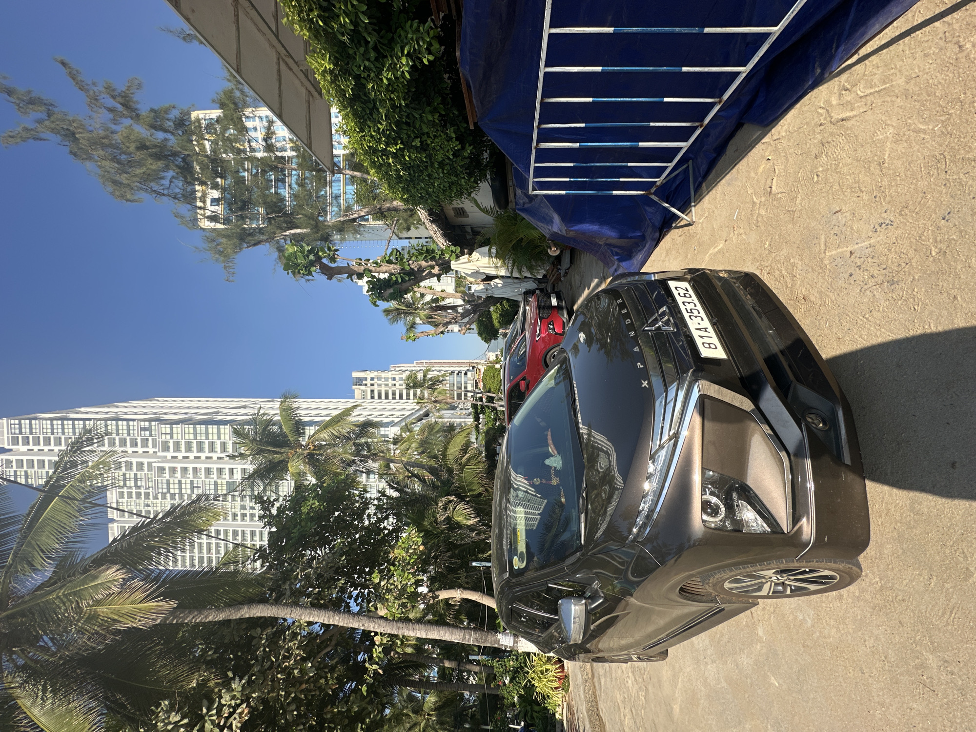 The image captures a street scene in Nha Trang, Vietnam, likely near a beach or resort area, given the presence of palm trees and tall buildings that resemble hotels or residential complexes. In the foreground, a dark brown Mitsubishi Xpander is parked on a paved surface, its license plate reading "81A-353.62". To the right of the car, a blue tarp is partially covering some materials, bordered by a white and blue metal fence. Further down the street, another car, a red SUV, is parked. The background is dominated by several multi-story buildings, some of which appear to be under construction or renovation, suggested by the blue tarp. The sky is clear and blue, indicating a sunny day. The overall impression is a coastal urban environment.