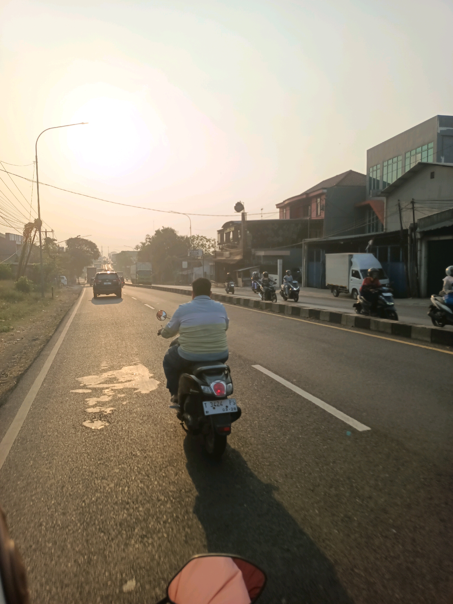 The image captures a street scene in Karawang, Indonesia, during what appears to be late afternoon or early morning, as indicated by the warm, low-angled sunlight creating long shadows and a hazy sky. The perspective is from someone riding a scooter, looking forward down a two-lane asphalt road.
In the foreground, the rear of a scooter is visible, with a portion of the passenger's seat and handlebar. The main subject of the image is a person riding a dark-colored scooter in the right lane, directly in front of the viewer. This rider is wearing a blue and yellow horizontally striped long-sleeved shirt and dark pants. The scooter has a license plate that is partially visible, reading "T 3424 P".
The road is relatively wide, with a white line marking the center and another white line on the right edge of the lane. There are several patches of water or wetness on the road surface, reflecting the sunlight.
On the right side of the road, there is a sidewalk area with a low concrete barrier. Beyond the sidewalk, several buildings are visible. These include a modern-looking building with a grey facade and large glass windows, a darker building with red accents, and other structures suggesting a commercial or mixed-use area. Several other scooters and motorcycles are also present on the road, some in the right lane, some in the left lane, and some parked or moving on the right side. One white delivery truck is also visible parked or moving slowly.
On the left side of the road, there is a grassy verge with scattered trees and vegetation. A prominent street lamp with wires attached to it stands on this side, and further down the road, more vehicles, including cars and trucks, are visible, creating a sense of moderate traffic. The overall impression is a typical urban or suburban street in Indonesia, with active local transportation and commercial activity.