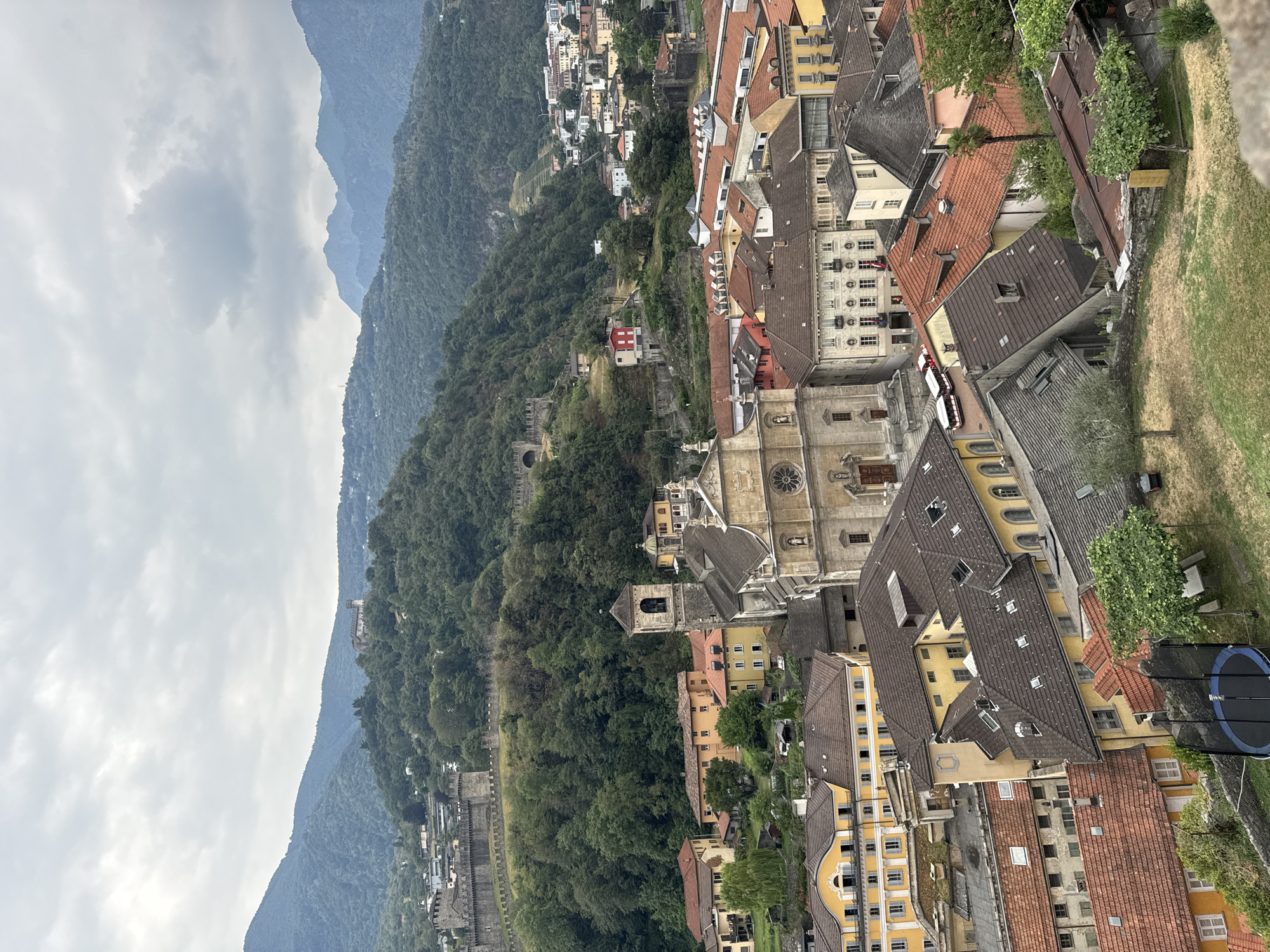 This is a high-angle, medium-shot photograph of the city of Bellinzona, Switzerland, focusing on its historic center and the surrounding mountainous terrain. The image is captured on an overcast day, with a muted color palette dominated by greens, browns, and grays.
In the foreground, a cluster of buildings with red and brown tiled roofs are visible. These structures exhibit a variety of architectural styles, with some featuring yellow facades and arched windows. A prominent church with a bell tower stands out in the mid-ground, its stone facade and steeple suggesting historical significance. The church is flanked by other buildings of varying heights and colors.
Behind the city, a lush green mountainside rises steeply, covered in dense forest. The slopes are dotted with more buildings, and in the upper left and upper center of the frame, imposing stone fortifications and castle walls are visible, indicating the presence of historical castles that are characteristic of Bellinzona. These fortifications snake along the hillsides, a testament to the city's strategic past.
In the distance, the peaks of larger mountains can be seen, their forms softened by the cloudy sky. A few vehicles, including a small red bus and what appear to be tourist trains, are present on the streets below, suggesting moderate activity within the city. The overall scene depicts a charming, historical European city nestled within a dramatic natural landscape.