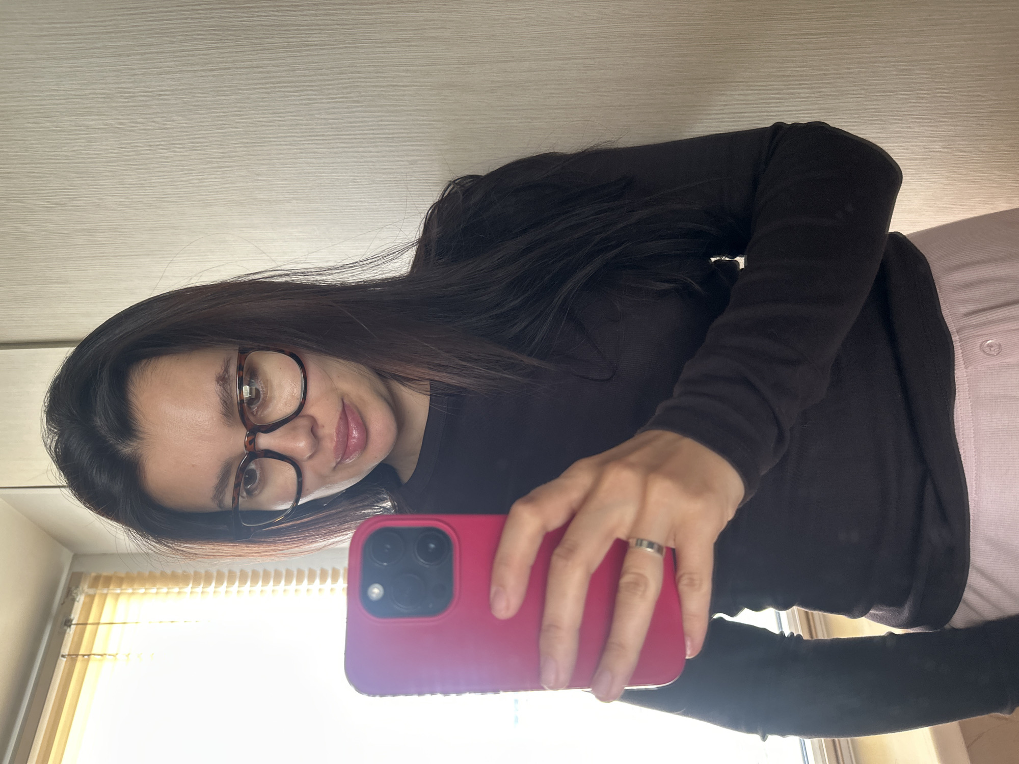 A woman is captured in a mirror selfie, looking directly into the camera with a neutral to slightly smiling expression. She has long, dark hair and wears dark-rimmed, possibly tortoiseshell, glasses. Her lips appear glossy. She is dressed in a dark, long-sleeved top, likely black or dark brown, and light-colored bottoms, possibly pink or light grey sweatpants.
In her right hand, she holds a bright pink smartphone with a visible multi-lens camera module, using it to take the photograph. A silver band-style ring is visible on her right ring finger.
The background suggests an indoor setting, with a light-colored wall or wardrobe door featuring subtle horizontal textures behind her. To the left side of the frame, a window with light-colored blinds is visible, allowing bright daylight to illuminate the scene. The presence of natural light indicates it is daytime. There is no visible text in the image.