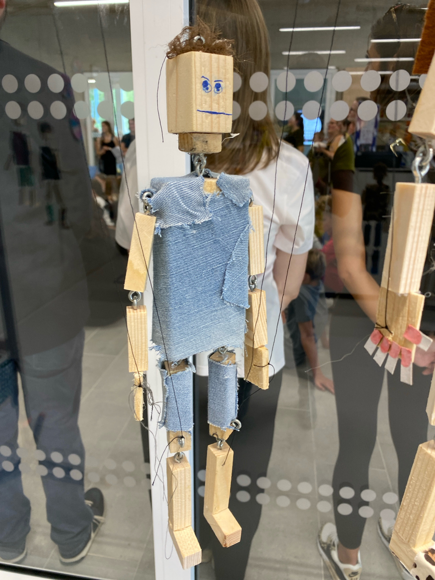A close-up, medium shot captures a handmade wooden marionette with a denim outfit, suspended by strings in what appears to be a public indoor space. The marionette's head is a simple wooden block with blue painted eyes and a drawn-on mouth. Its body and limbs are constructed from wooden pieces and connected with metal rings, all draped in torn denim fabric. The background, visible through a reflective surface, shows a blurred scene of people in what could be a museum, gallery, or exhibition space, with a woman in a white shirt and black leggings partially obscuring the marionette. The floor is tiled, and the overall lighting suggests an indoor environment. There is no specific indication of the location being in Horhausen (Westerwald), Germany.
