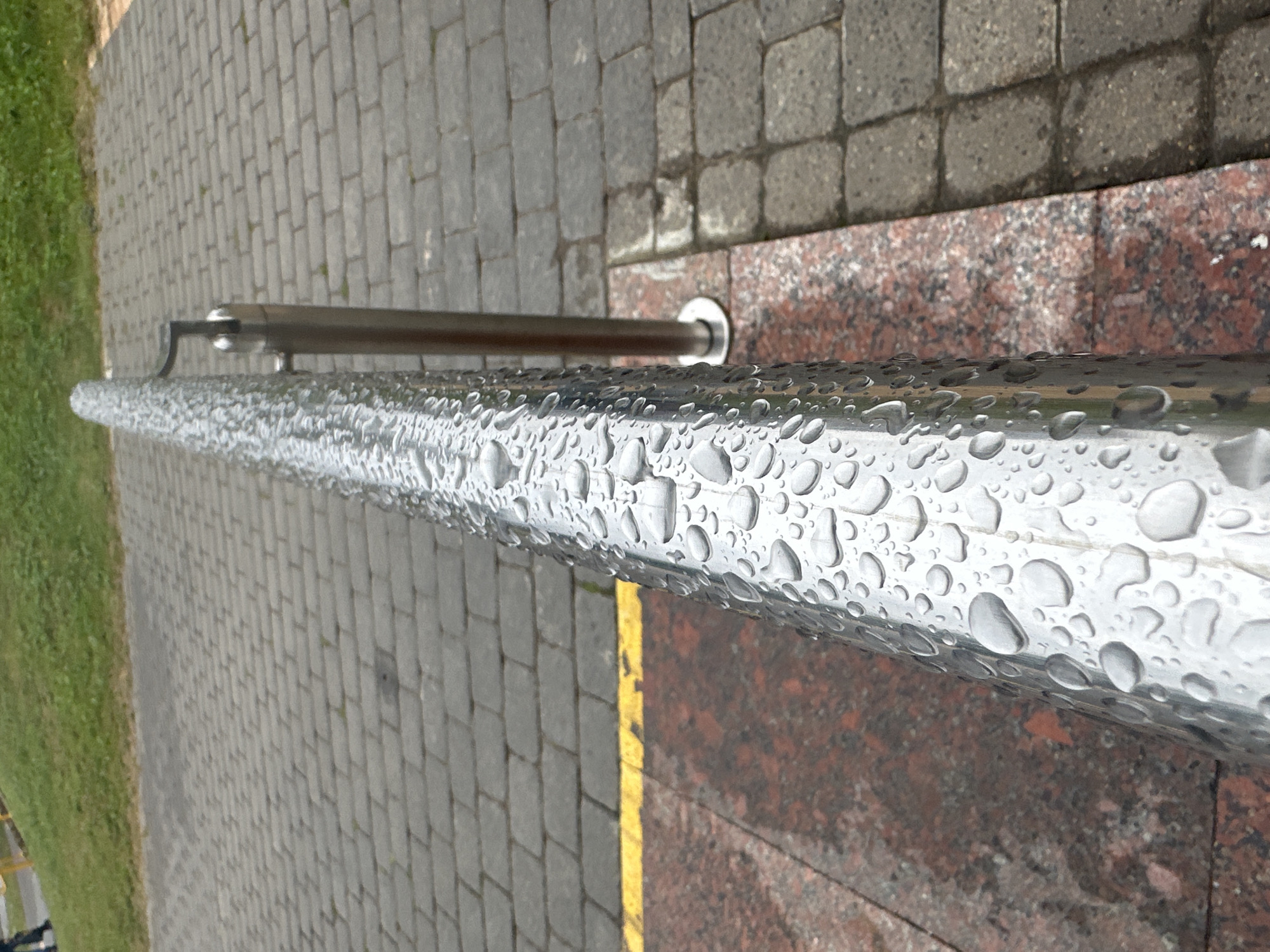 The image shows a close-up of a wet metal handrail in an outdoor urban setting. The handrail is cylindrical and covered in numerous water droplets, indicating recent rain. It is supported by a vertical metal pole. The background consists of a paved walkway made of gray interlocking stones. To the left of the handrail, there is a distinct yellow line bordering a reddish-brown granite surface. Beyond this, a patch of green grass is visible. In the far upper left corner, the legs of a person can be seen wearing dark pants. The scene appears to be a public area, likely a pathway or steps, with the paving stones suggesting a pedestrian zone in Minsk, Belarus.