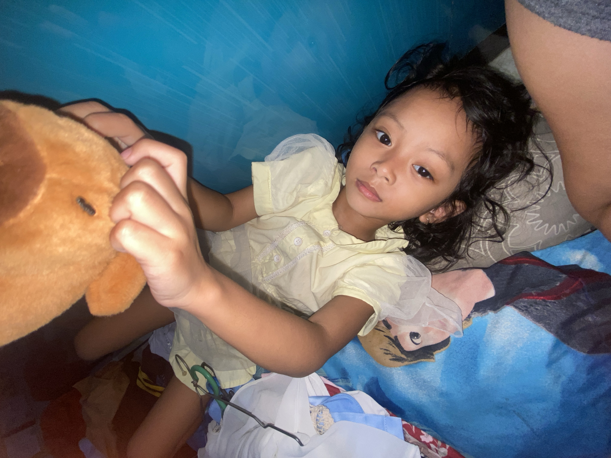 A young child, likely a girl, with dark, curly hair and brown eyes, is captured lying on her back, looking directly at the camera with a calm, neutral expression. She is wearing a light yellow short-sleeved top adorned with white lace trim and sheer, puffy sleeves. Her right hand gently holds a brown, plush stuffed animal, possibly a dog or bear.
The child is positioned indoors, resting on what appears to be a bed or floor. Her head is supported by a grey pillow featuring a white geometric pattern. Beneath her and to her right is a vibrant blue blanket, decorated with a depiction of the animated character Naruto. Around her lower body and to her left, various items of clothing or fabric in shades of white, blue, and other patterns are visible. A metal clothes hanger, with green and silver components, lies near her left elbow.
The background directly above her head is a solid blue surface, which could be an indoor wall, a tent fabric, or a large draped sheet. The lighting suggests an indoor setting, possibly with artificial light, creating soft shadows. There is no visible text in the image. The overall scene depicts a relaxed moment in a personal, somewhat cluttered, indoor space.