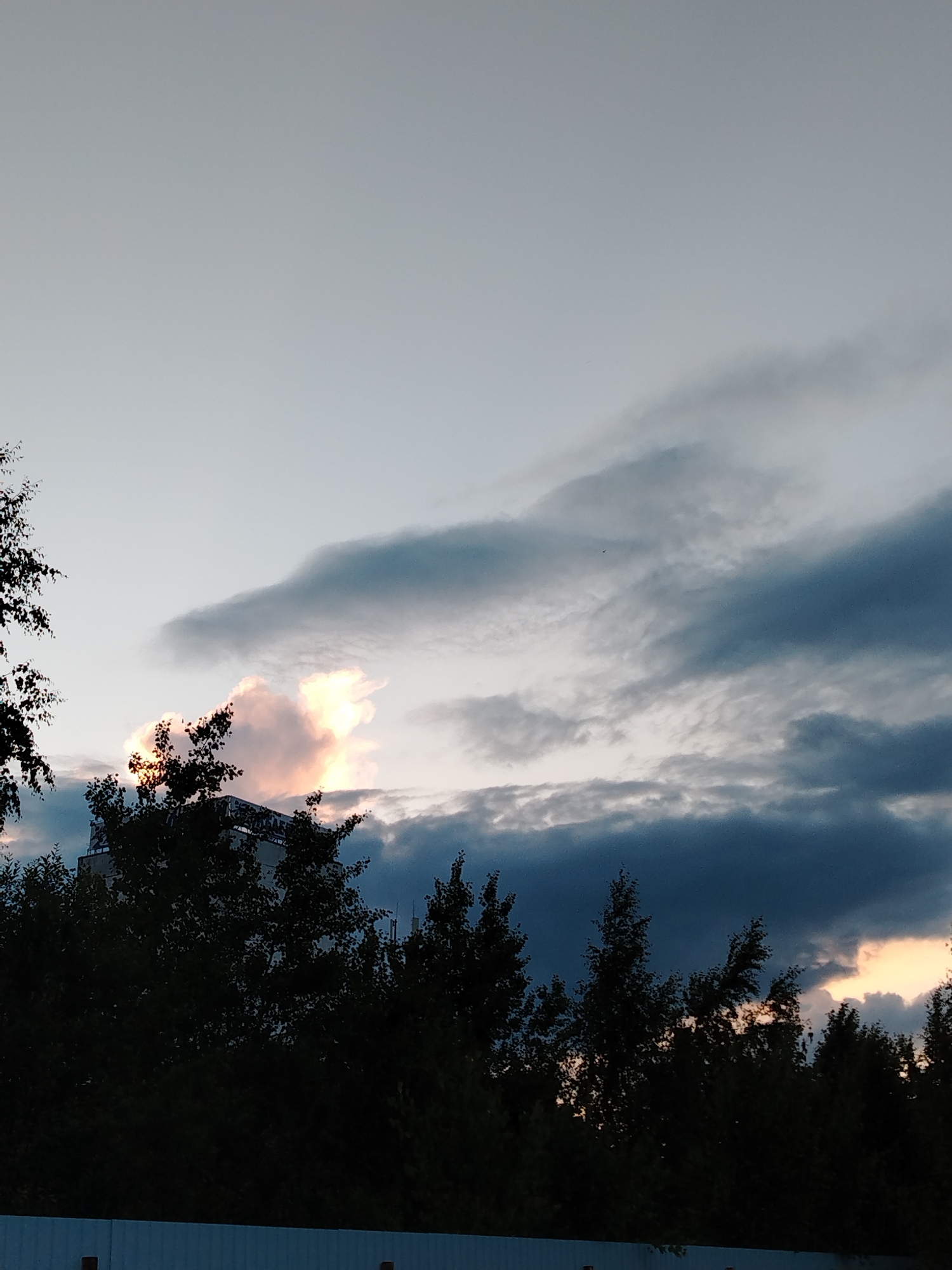 A wide-angle shot captures a dusky sky over a treeline in Zavolzhye, Russia. The sky is a gradient of light gray and deep blue, with dramatic, dark clouds dominating the upper right portion of the frame. A bright, illuminated cloud, tinged with orange and pink, is visible in the middle of the frame, suggesting the presence of the setting sun. Silhouetted against the sky are the dark shapes of trees, some with visible branches and foliage. Partially visible behind the trees is the top of a building with a light-colored facade, possibly a multi-story structure. In the foreground, a portion of a white corrugated fence runs horizontally across the bottom of the image. The scene appears to be a quiet, naturalistic view of the transition from day to night in an urban fringe environment.