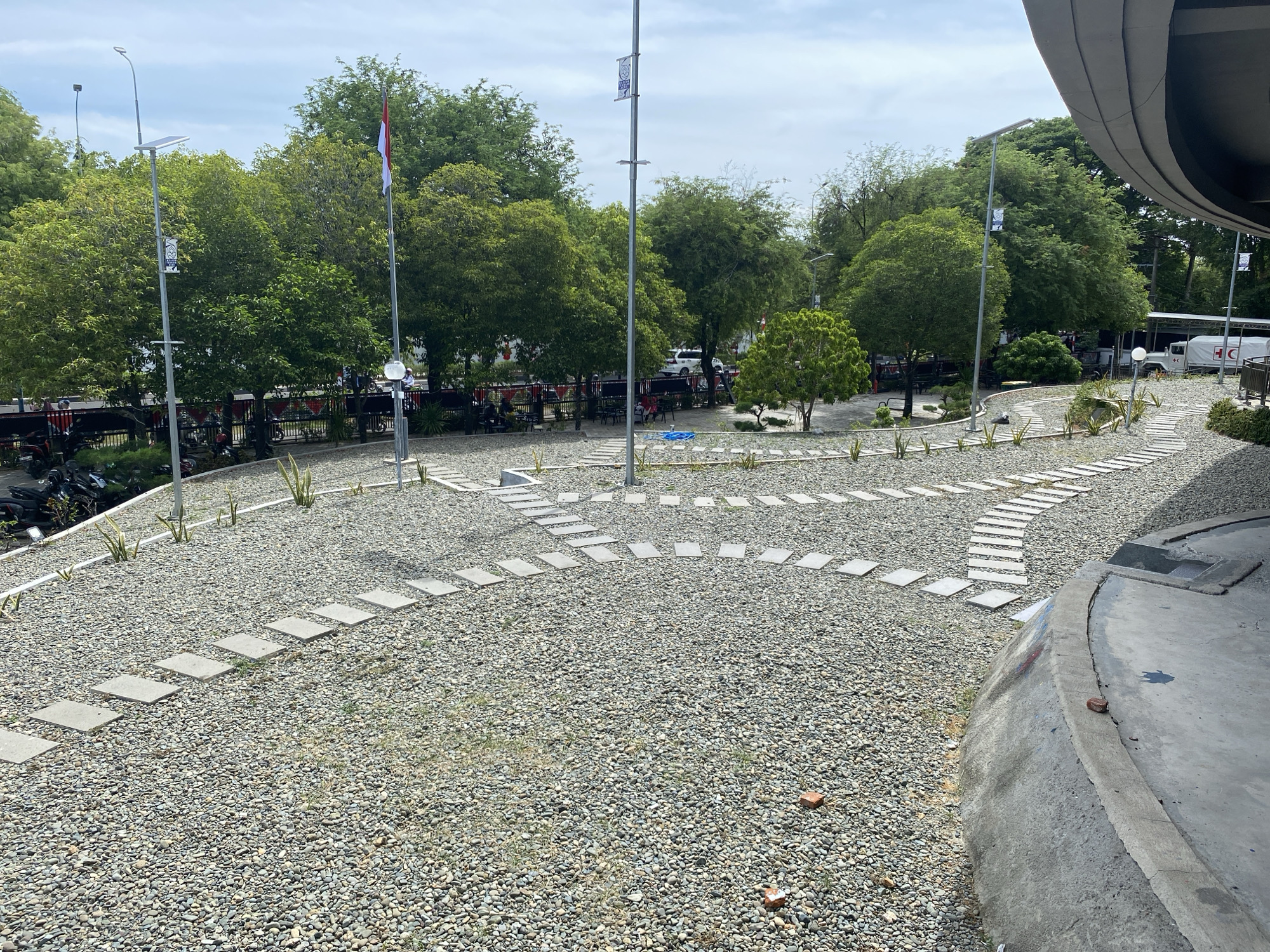 This is a medium-angle, eye-level shot of an outdoor area in Banda Aceh, Indonesia. The foreground is dominated by a pathway of square, light-colored pavers set into a bed of small, grey pebbles. The path curves gently to the right and then curves back to the left. Sparse, thin green plants are strategically placed between some of the pavers. Further back, a line of trees with lush green foliage forms a backdrop. A flagpole with a red and white flag is visible on the left side of the image, behind the trees. A modern, grey, curved structure is visible in the upper right corner of the image. In the middle ground, there appears to be a shaded seating area with tables and chairs, and several people are seated. In the background, there is a fence with red and white banners, and further back, a white truck with red markings is parked. The sky is overcast with white clouds. The scene appears to be a public space, possibly a park or plaza, given the presence of seating, pathways, and landscaping.
