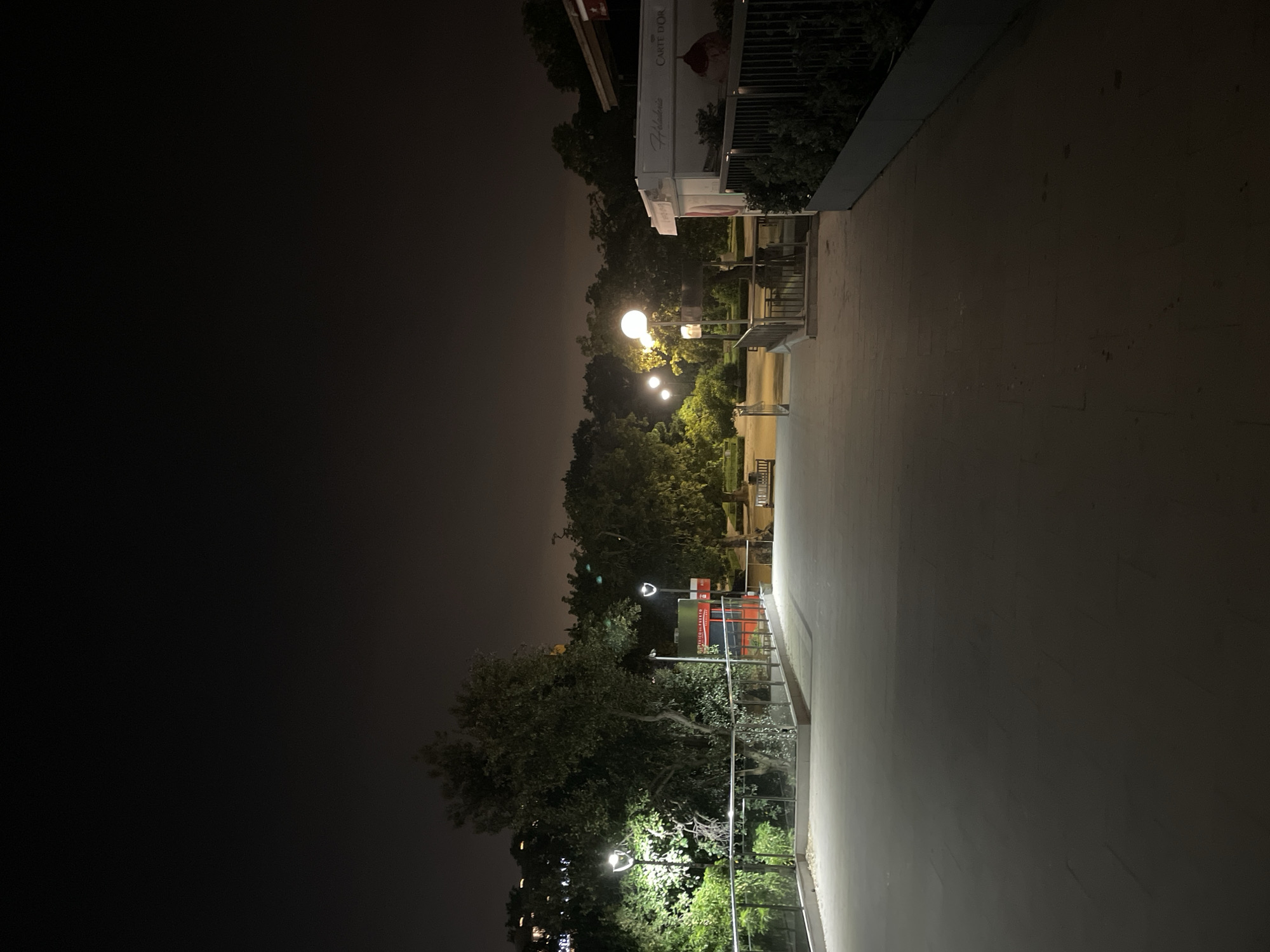 The image depicts a nighttime scene along a walkway in what appears to be a park or public space in Barcelona, Spain. The viewpoint is looking down the path, with a structure, possibly a kiosk or small shop, visible on the right side near the top of the image. Trees and shrubbery line the side of the path with lighting fixtures illuminating the area. The sky is dark, with some light visible through the trees suggesting the presence of buildings in the distance. The path itself is made of a light-colored material, contrasting with the dark surroundings. The image is oriented such that the sky is on the left and the walkway leads off to the right.