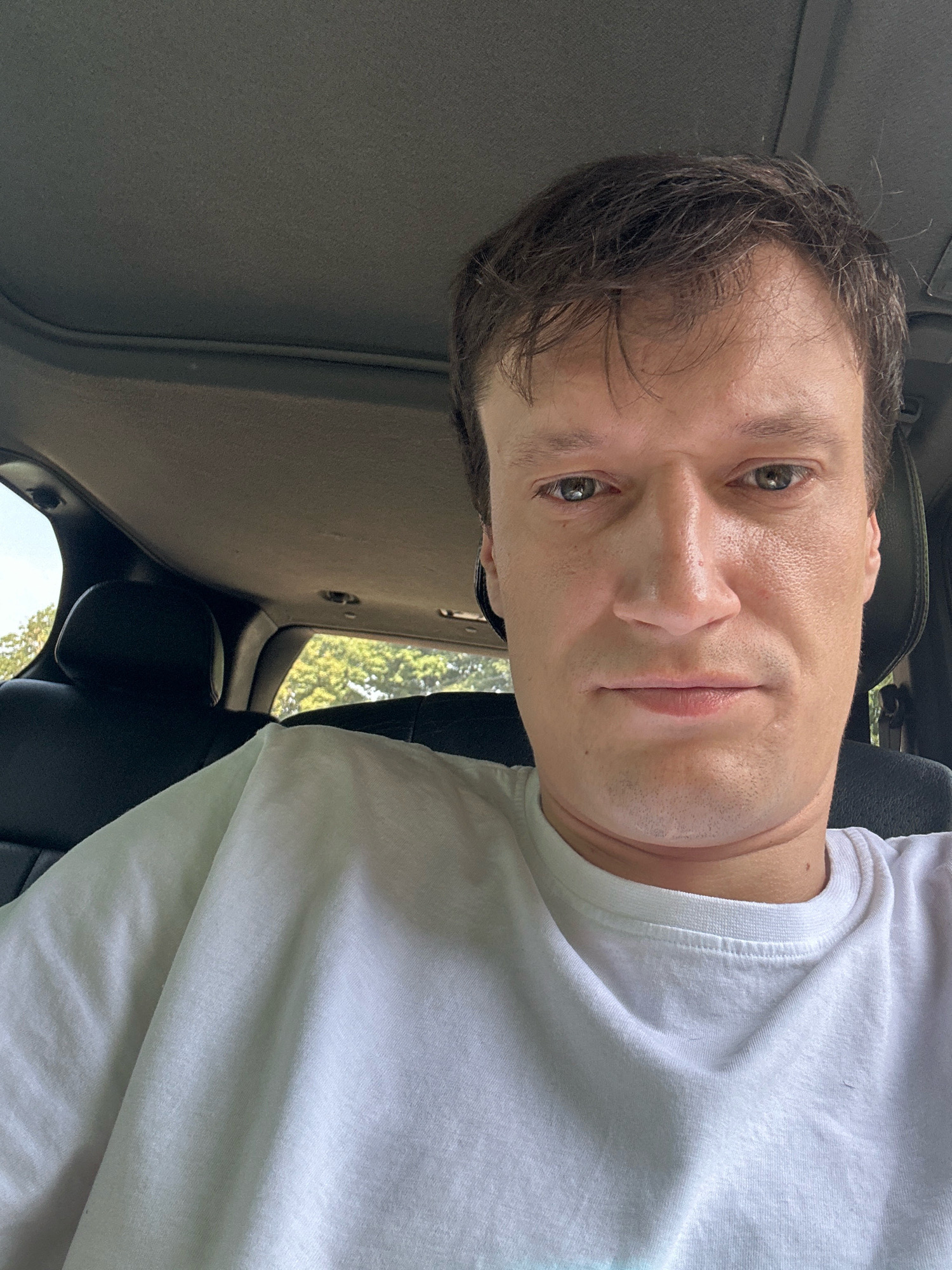 A man with brown hair and hazel eyes is pictured from the chest up, looking directly at the camera. He is wearing a white t-shirt and appears to be inside a vehicle, with the interior of the car visible in the background, including the sunroof and seats. The exterior of the car shows trees and sky through the windows. The overall lighting suggests it is daytime. There is no discernible indication of the specific location within the city of Kamianets-Podilskyi, Ukraine.