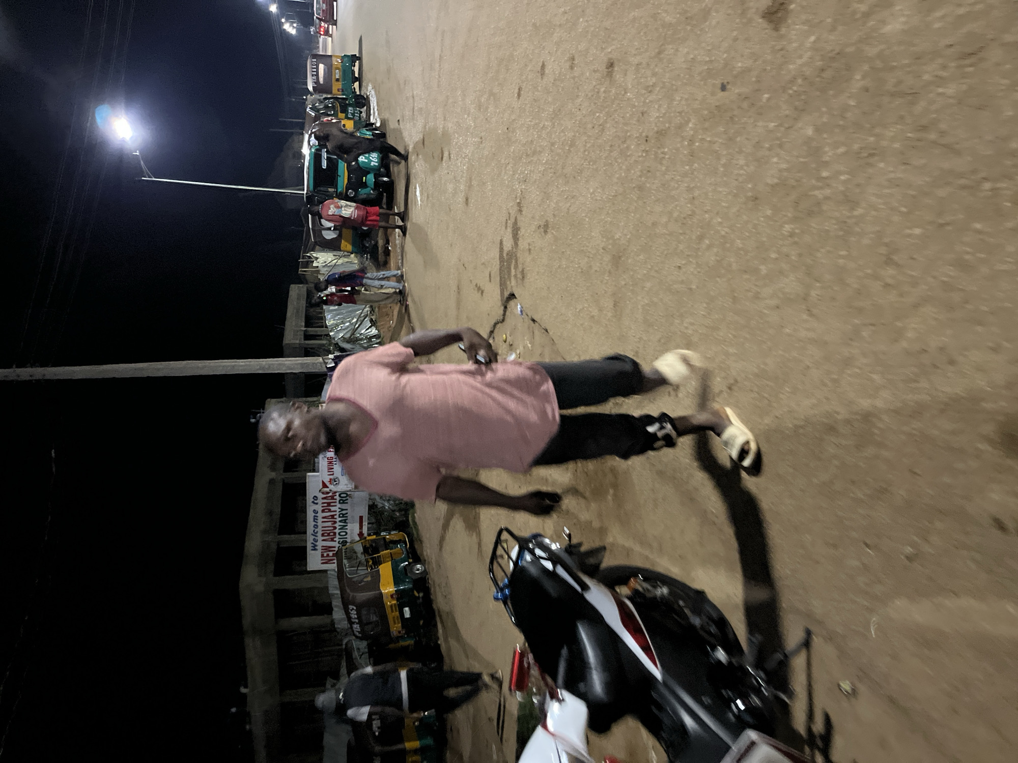 This nighttime image captures a street scene, likely in an urban or semi-urban area of Jos, Nigeria. The photograph is somewhat blurry due to low light or motion and is vertically oriented, showing a street with activity.
In the foreground and midground, a man wearing a pink t-shirt, dark trousers, and light-colored sandals walks on the road, looking towards the left side of the frame. He appears to be holding a small object in his hand. A black and white motorcycle is partially visible in the bottom right corner.
Along the left side of the street, several motorized tricycles, commonly known as Keke Napep, are parked. These tricycles, seen in various colors like green, yellow, and brown, serve as local public transport. Some display visible text such as "PA-1469", "PTA 0154", "7646", and "PTA-11101". Several other individuals are gathered near these vehicles.
A tall street light illuminates a section of the area from the left, highlighting the road and parts of the parked vehicles. Near the tricycles, some makeshift structures or stalls are visible. A white sign with red and black text partially reads, "Welcome to NEW ABUJA PHAS" and "LIVING SIONARY RO", suggesting a welcome sign to "New Abuja Phase" and a "Missionary Road" or similar. The dark sky confirms the time of day as night. The ground appears to be a light-colored asphalt or dirt road. The scene portrays a typical evening atmosphere of commerce and transport in a Nigerian locality.