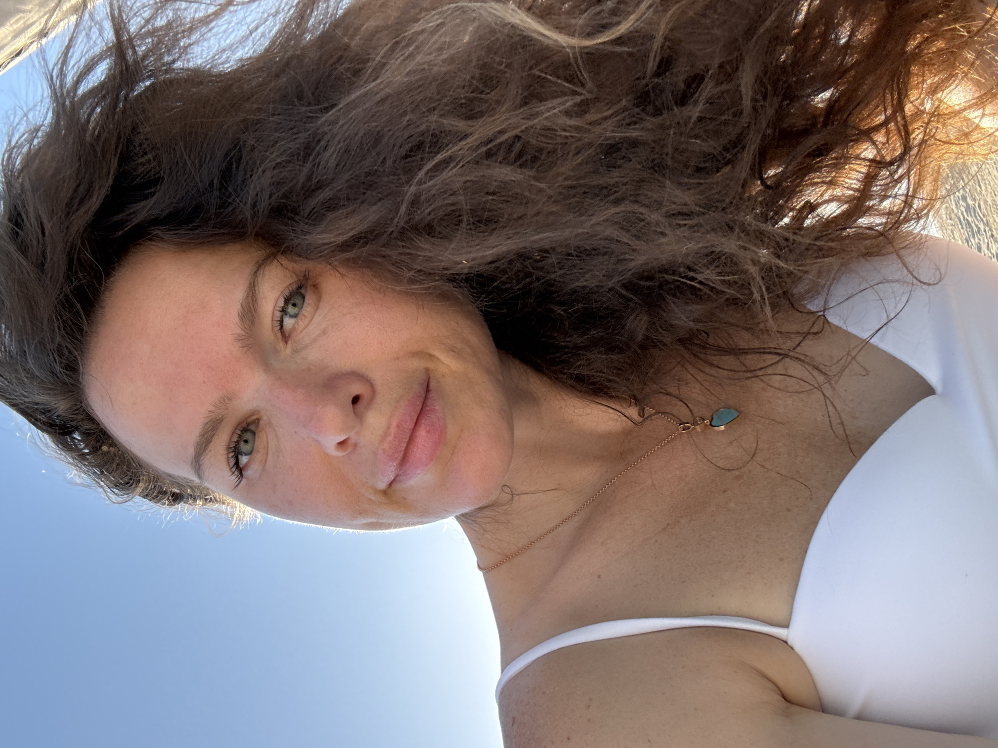 A close-up selfie captures a woman with long, dark, wavy hair against a clear blue sky. She is wearing a white swimsuit and a delicate gold necklace with a blue pendant. The lighting suggests it is daytime, possibly outdoors, with sunlight highlighting her features and hair. Her expression is relaxed and she is looking towards the camera. The background is simple, with no discernible landmarks to indicate a specific city or location.