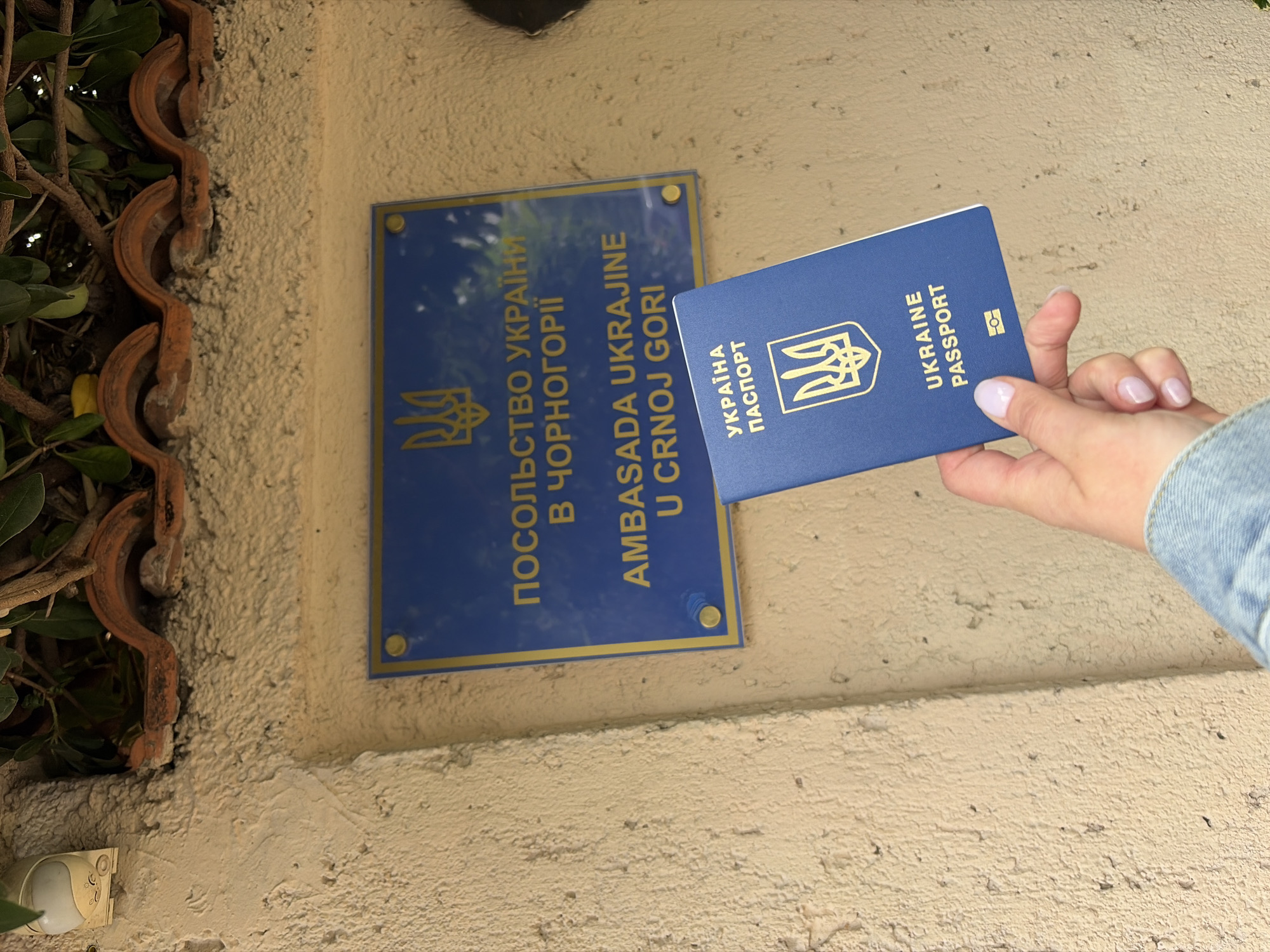The image shows an embassy sign and a passport held by a person. The sign reads "ПОСОЛЬСТВО УКРАЇНИ В ЧОРНОГОРІЇ AMBASADA UKRAJINE U CRNOJ GORI," which translates to "Embassy of Ukraine in Montenegro." The sign is blue with yellow lettering and is mounted on a beige textured wall. A Ukrainian passport, blue with the Ukrainian coat of arms and the words "UKRAINE PASSPORT," is held in front of the sign by a hand. The person's hand is pale-skinned with manicured nails and wearing a denim jacket. To the left of the sign, there are decorative terracotta tiles and greenery. The setting appears to be outdoors. The location is likely near the Ukrainian Embassy in Podgorica, Montenegro, given the signage.