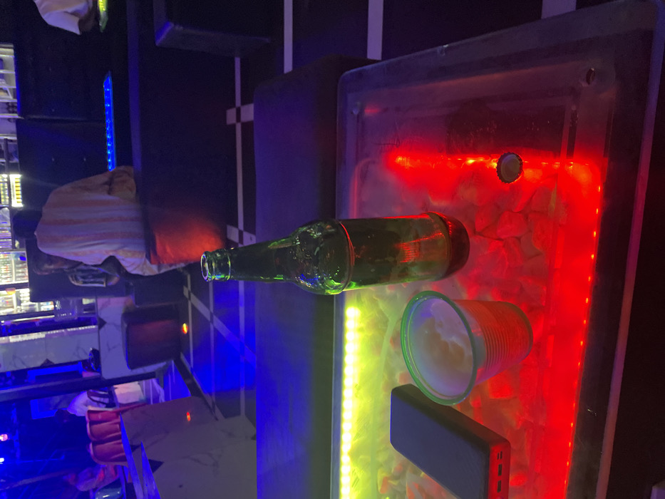An empty green glass bottle lies horizontally on a table, alongside a translucent plastic cup filled with a white substance. A dark rectangular object, possibly a power bank, is near the cup. The scene is illuminated by red, yellow, and blue neon lights. The table appears to be made of glass or clear plastic, resting atop small white stones. In the background, a person is seated in a booth.