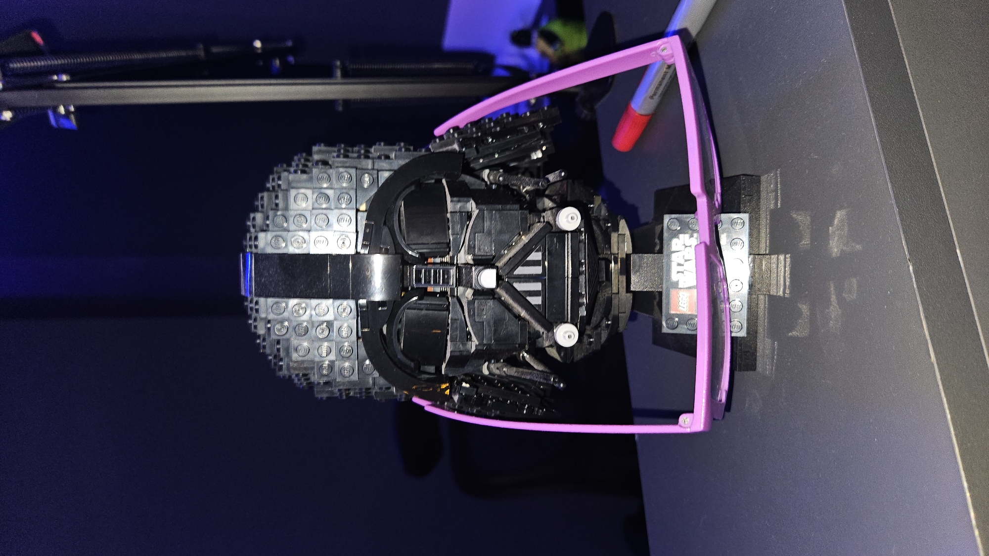 In an indoor environment with dark blue walls, a LEGO Darth Vader helmet is prominently displayed on a black stand. The helmet is adorned with a pair of pink-rimmed glasses perched on its face. To the right of the helmet, a red-tipped permanent marker lies on a dark grey surface, likely a desk or table. The scene is dimly lit, with a faint purple light emanating from the background. A black microphone stand is visible in the far right background.