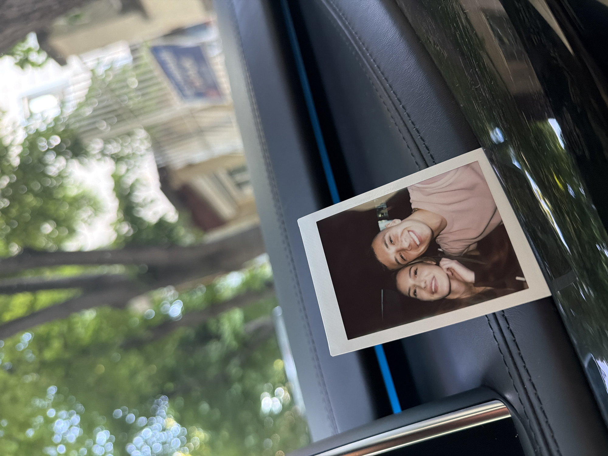 A Polaroid photograph of two women, smiling and posing, is affixed to the interior of a car. The car's interior is leather with blue accents. Through the car window, an out-of-focus cityscape is visible. The location is Plovdiv, Bulgaria.