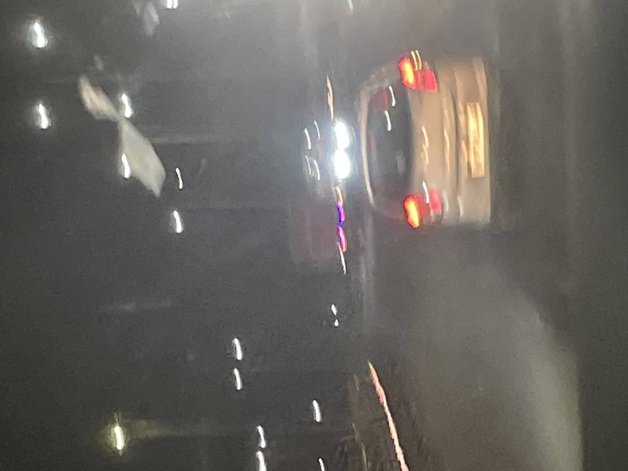 The image appears to be a photograph taken at night, possibly from inside a vehicle, given the motion blur and perspective. The scene is dark, with numerous lights that are blurred and streaky due to movement.
The primary subject appears to be a light-colored vehicle with its red taillights illuminated, seen from the rear. The vehicle is slightly tilted. In the foreground and scattered throughout the image are various bright, blurry lights, some appearing as streaks and others as more concentrated orbs. Some of these lights are white, while others have a reddish or yellowish hue, consistent with vehicle headlights or streetlights. There are also some indistinct shapes that might be a person or object, partially obscured and blurred. In the top left, there is a white, amorphous shape that is also blurred, possibly a piece of cloth or debris.
The overall impression is that of a moving scene at night, characterized by artificial lights and a lack of clear detail. There are no visible text elements in the image. The context provided, "B Camp, India," suggests the location, but this information cannot be visually verified from the image itself. The time of day is clearly nighttime, and the weather is not discernible. There are no discernible emotions conveyed due to the lack of clear subject detail and the abstract nature of the light streaks.