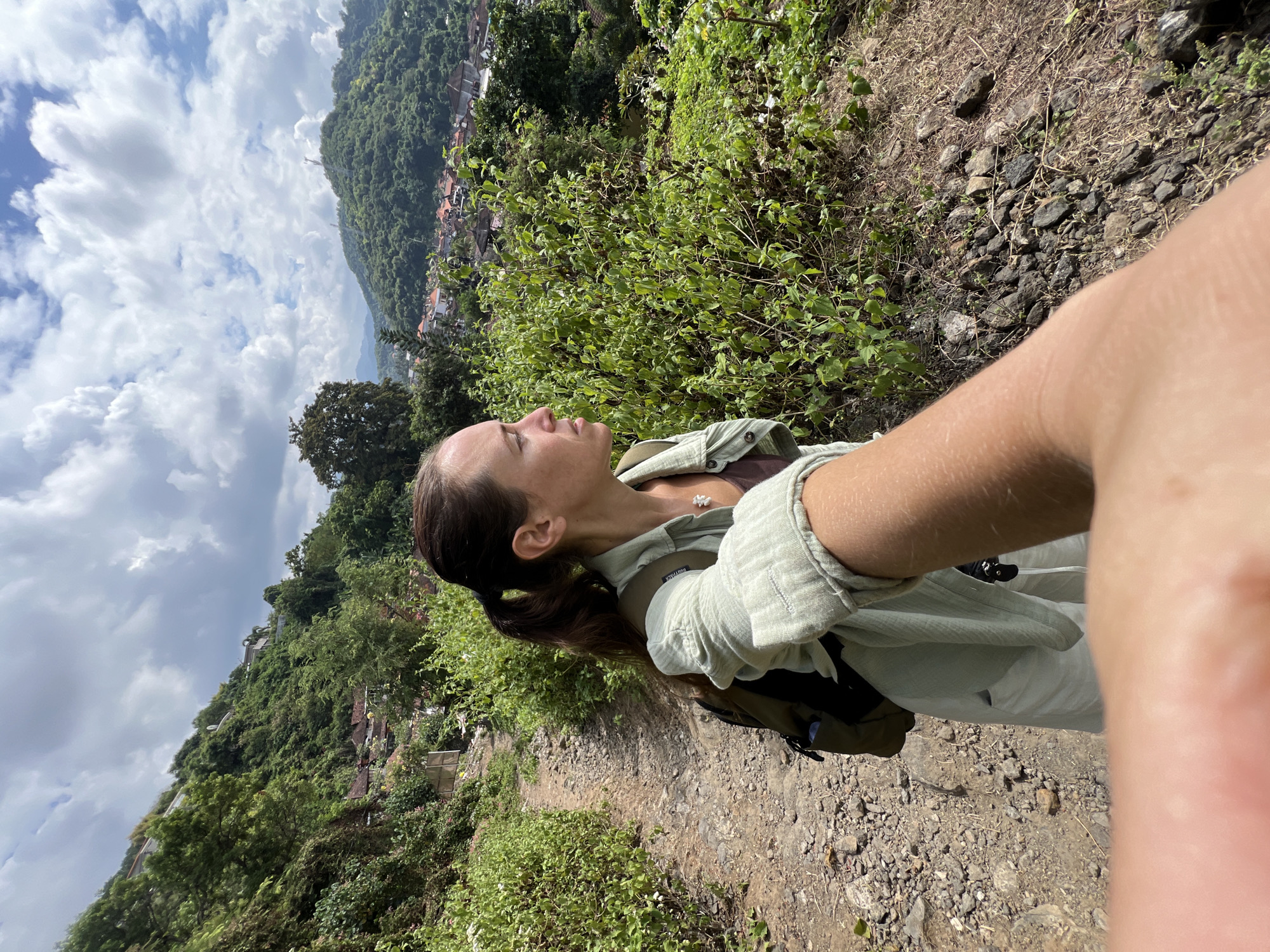 An adult female with brown hair pulled back in a ponytail is captured in a selfie from the chest up, looking to her right. She wears a light green button-up shirt over a brown tank top and has a backpack on. The woman stands on a dirt path surrounded by lush green vegetation. In the background, a village with houses is nestled among rolling, tree-covered hills under a partly cloudy sky. The scene is identified as being in Manggis, Indonesia.
