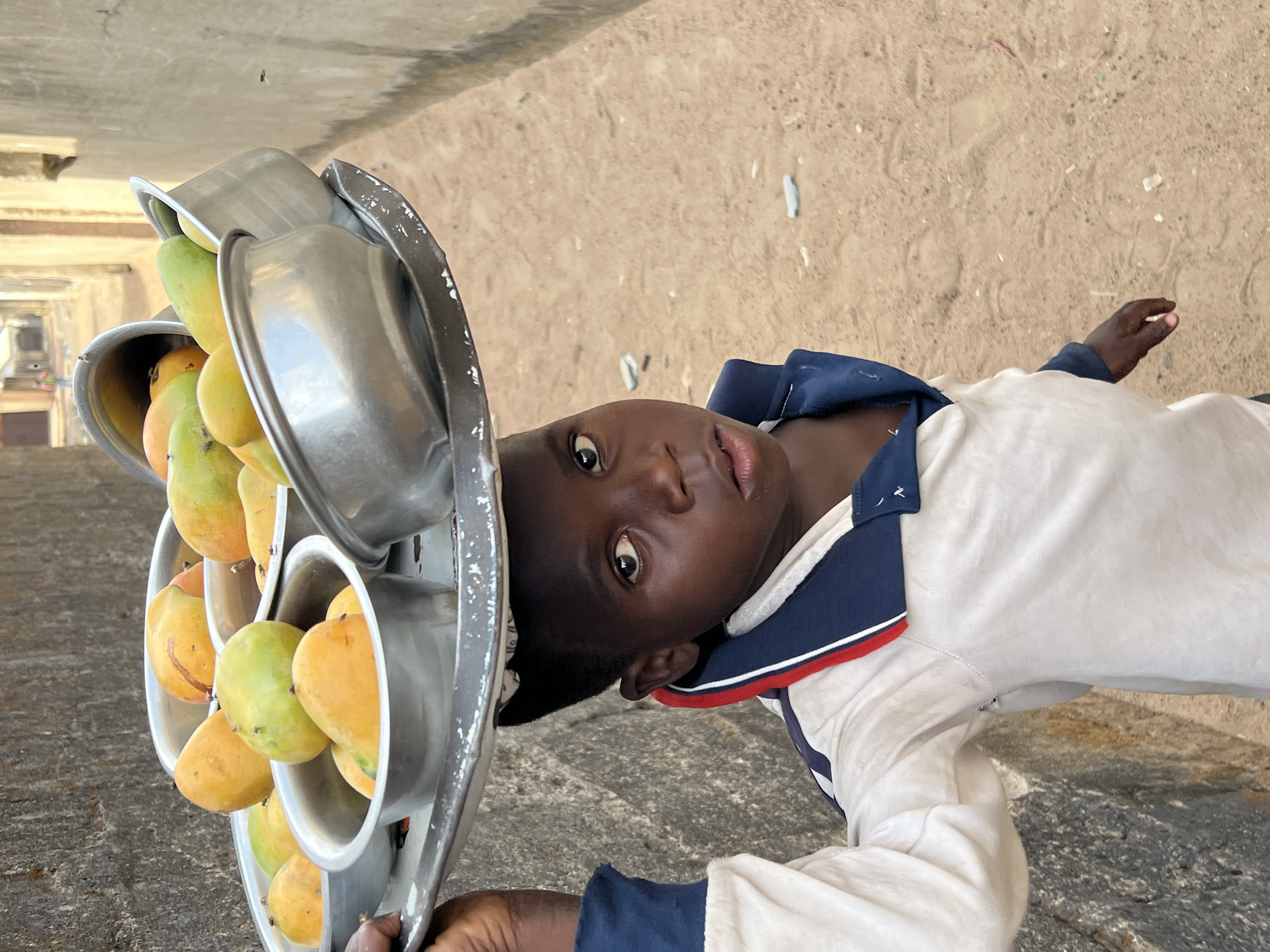 A young person with dark skin is carrying a metal container filled with mangoes, which rests on their head. The mangoes are yellow and green. The person is wearing a white shirt with a navy blue and red collar. The background is a sandy color. This image was likely taken in Minna, Nigeria.