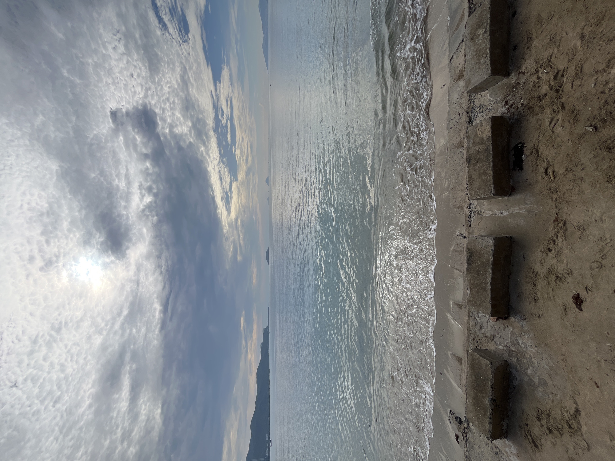 The image depicts a coastal scene, possibly a beach, oriented vertically. The sky occupies the upper portion of the frame, showing a mix of bright and dark clouds. The sun's light filters through, creating a bright patch. Below the sky is the sea, stretching towards the horizon. The water appears relatively calm, with slight ripples and reflections. A coastline or landmass is visible in the distance. The foreground is dominated by a sandy area, along which are arranged concrete blocks or steps. The blocks are gray and appear weathered. Some are partially buried in the sand. The edge of the beach meets the water, with small waves lapping at the shore. The image may be depicting the city of Bac Nha Trang Ward, Vietnam. There are no people visible.
