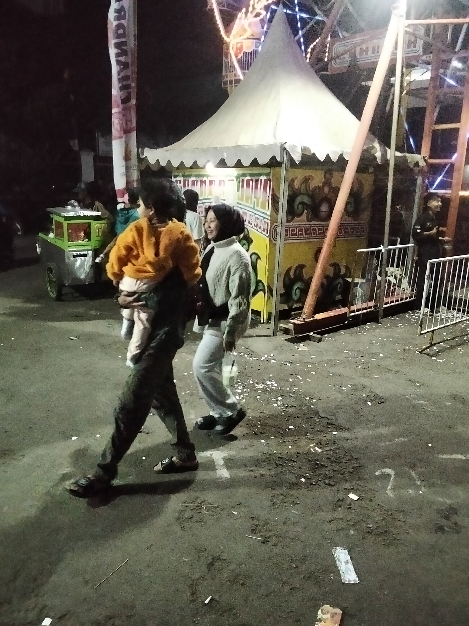 In Cisaat, Indonesia, a night scene unfolds at what appears to be a fairground. A man, seen from the knees down, walks left to right, carrying a child on his shoulders. The child is clad in an orange sweatshirt and pink pants, with their arms around the man's neck. Beside them, a woman in a light-colored outfit walks with her head tilted to the side, seemingly in conversation with the man. Behind them, a tent with white scalloped edges and a yellow backdrop stands, decorated with abstract patterns. A small food cart, green and silver, is visible to the left. The ground is a mixture of dark asphalt and scattered debris, illuminated by overhead lights and festive string lights from an unseen amusement ride. A white metal fence separates the walkway from another area.