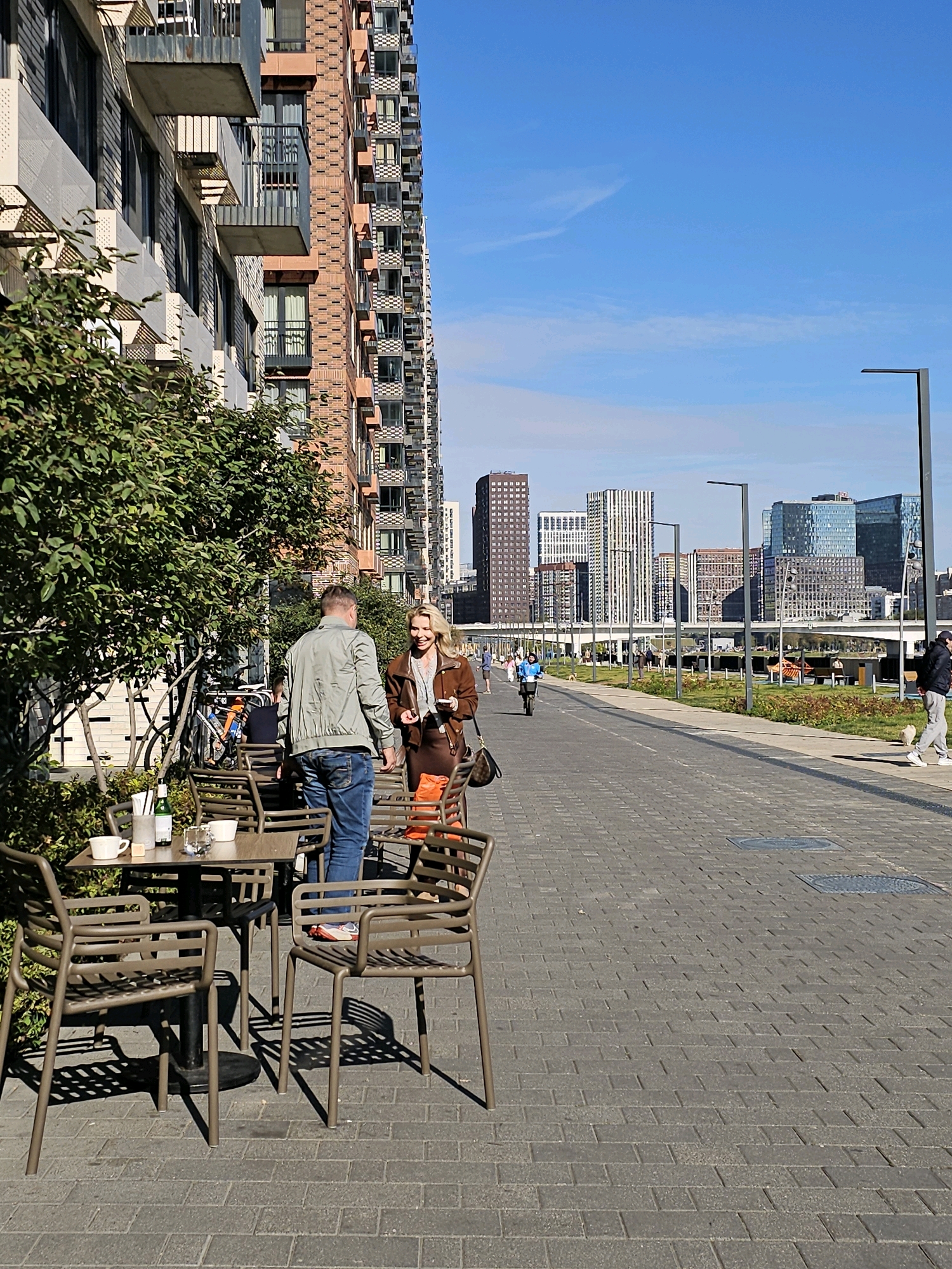 A man and a woman stand next to an outdoor cafe table on a brick-paved walkway in Moscow, Russia, near the Moscow River. The man, wearing a green jacket and blue jeans, faces away from the camera. The woman, in a brown jacket and skirt, faces the man and smiles. A table with white cups and a bottle of wine is visible. In the background, a modern residential building with balconies dominates the left side of the frame, while a cityscape of tall buildings and a bridge stretches across the horizon. The sky is clear and blue.