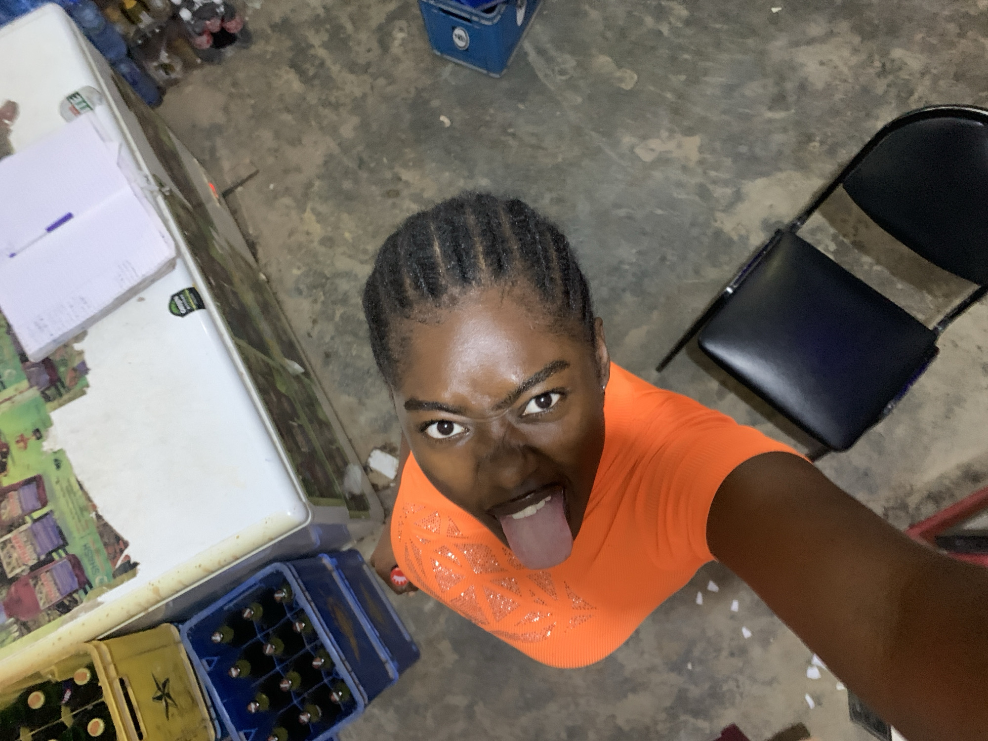A medium-angle shot looking upward at a person in an orange, short-sleeved shirt with an angry expression, sticking their tongue out. The person has dark skin and braided hair. Behind them is a gray, concrete-like floor, a folding chair with a black seat and back, a light-colored refrigerator with a decorative front, and several crates of beverages. A bottle of beverages sits near the front. The location is New Stadium, Nigeria.
