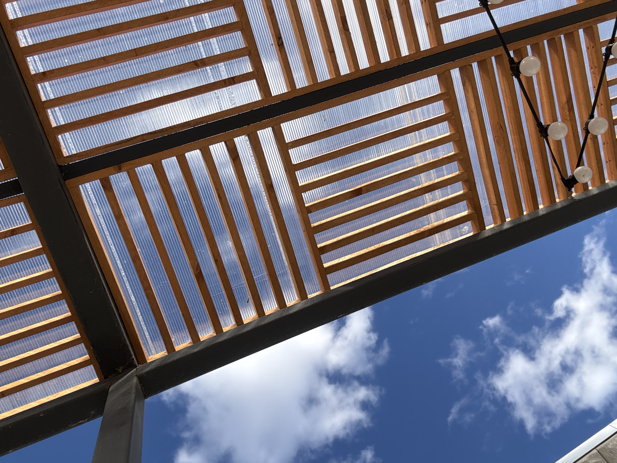 The image shows a view looking upwards at a wooden pergola structure with a clear corrugated roof covering. The pergola is supported by dark grey metal beams and posts. A string of light bulbs hangs from one corner of the structure. The background is a blue sky with scattered white clouds. The image suggests an outdoor space, likely a patio or garden area. The location is not identifiable from the image.