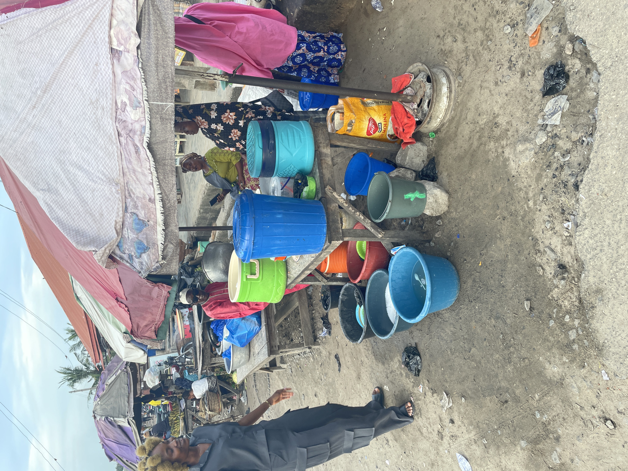 The scene depicts a street in Badagry, Nigeria. A market stall displays various colorful buckets, plastic containers, and cookware. Two women are standing behind the stall, one in a green top and another in a floral dress. A bag of "PRIMA FOODS MOLINA" is visible among the goods. A man in a dark suit and sandals stands in the foreground. The ground is cracked and littered with debris. Overhead, a makeshift awning provides shade. In the background, there are other stalls and pedestrian activity.