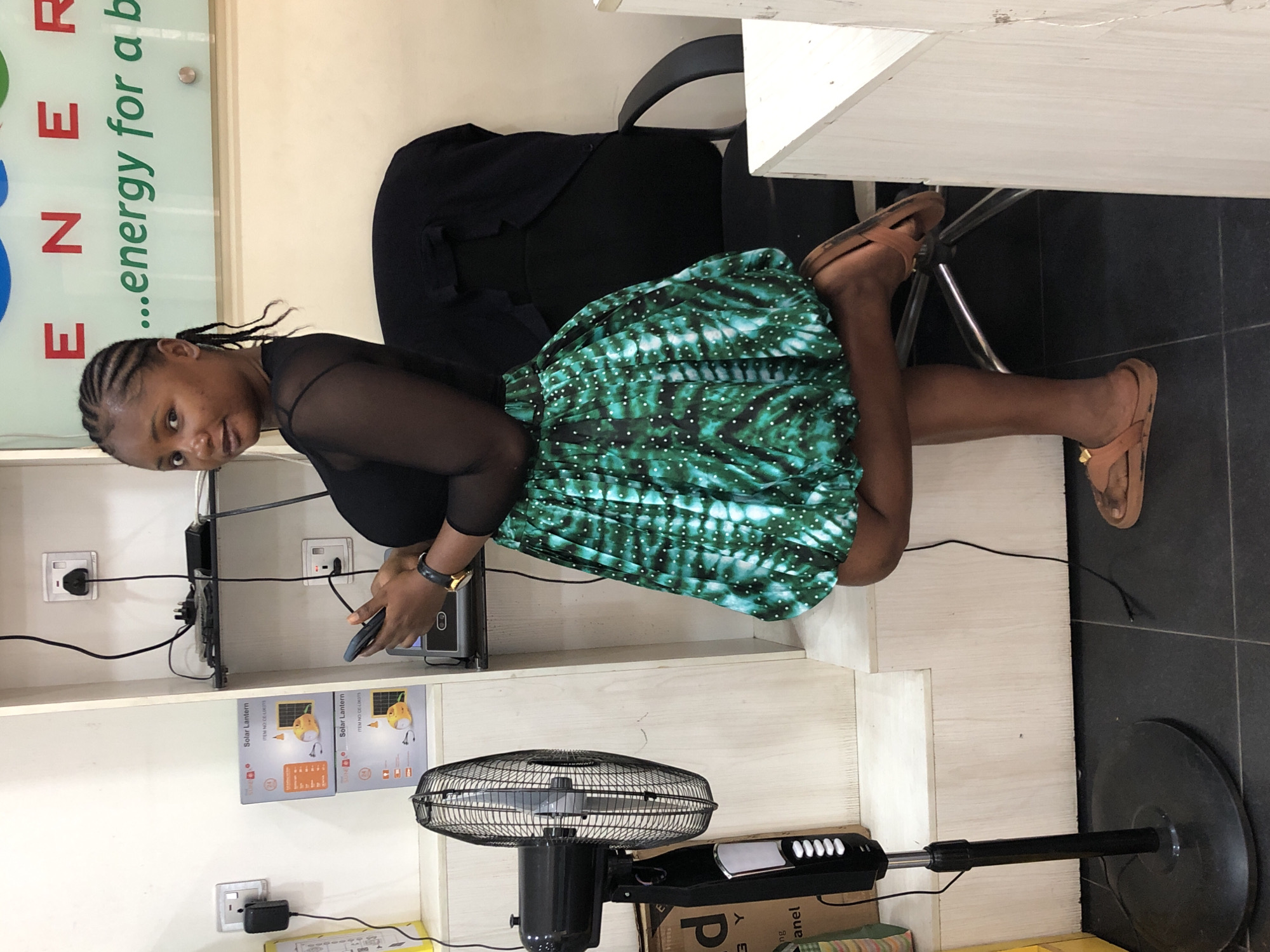 A young woman with braided hair, wearing a black sheer top and a green and white patterned skirt, stands with her body angled to the right, holding a smartphone. She is in an indoor setting, possibly a store or office, in Ikeja, Nigeria. To her left, a black standing fan is positioned on the floor. Behind her, shelves are visible, holding boxes labeled "Solar Lantern" and electrical outlets. A black office chair is behind her to the right. The background features a wall with a sign that reads "ENERGY ...energy for a b". The floor is tiled in a dark color.