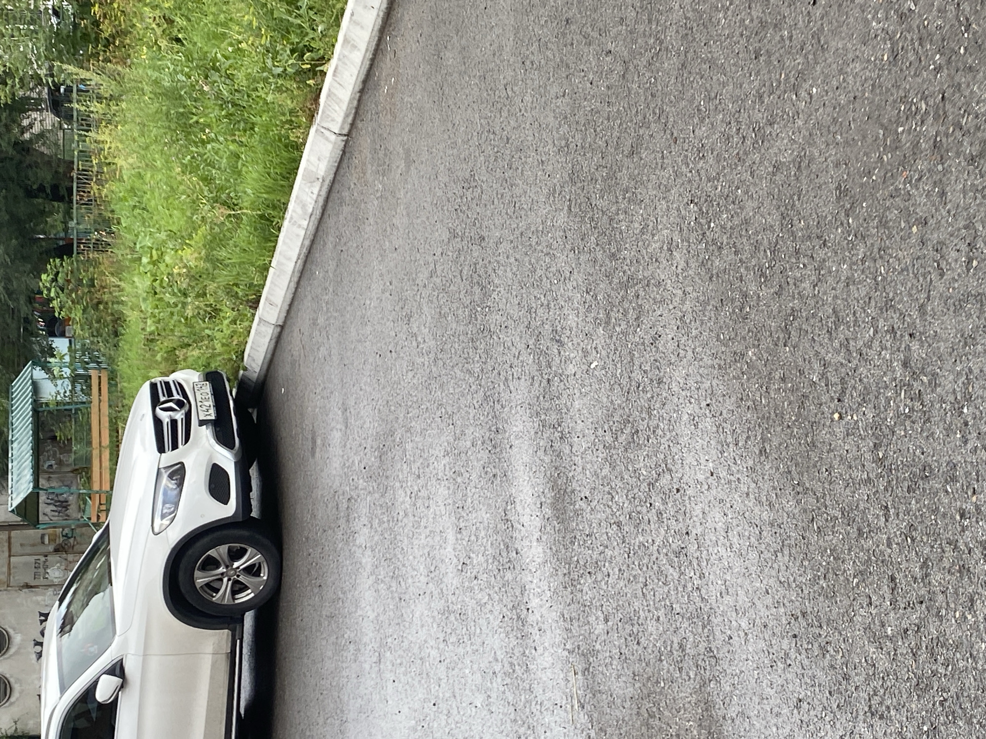 A white Mercedes-Benz GLC-Class SUV is parked at the side of an asphalt road. The car's front left wheel is turned slightly outward and is near a concrete curb. The SUV has a license plate that reads "X421EO 199". Behind the curb, there is overgrown green grass and some trees. In the background, there are parts of a building and a green metal fence. The scene appears to be in an urban or suburban environment.