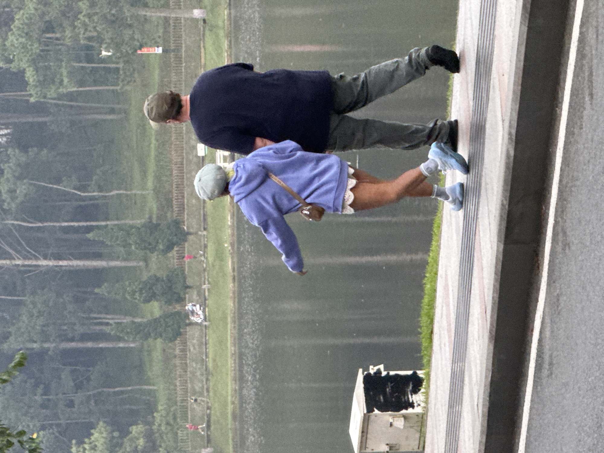 Two individuals, a woman and a man, are walking on a paved walkway next to a body of water. The woman, in the foreground and to the left, is wearing a light blue hooded jacket, a white lacy skirt, and light blue sneakers. She has a small brown crossbody bag. The man, to the right, is wearing a dark blue t-shirt, olive green cargo pants, a green baseball cap, and dark shoes. They are walking away from the camera, towards the right side of the frame. In the background, there is a body of water, a grassy embankment, and a line of trees. Further in the distance, there are more trees and a hazy, overcast sky. A utility box is present on the left side of the walkway. The location is identified as Phường Xuân Hương - Đà Lạt, Vietnam.