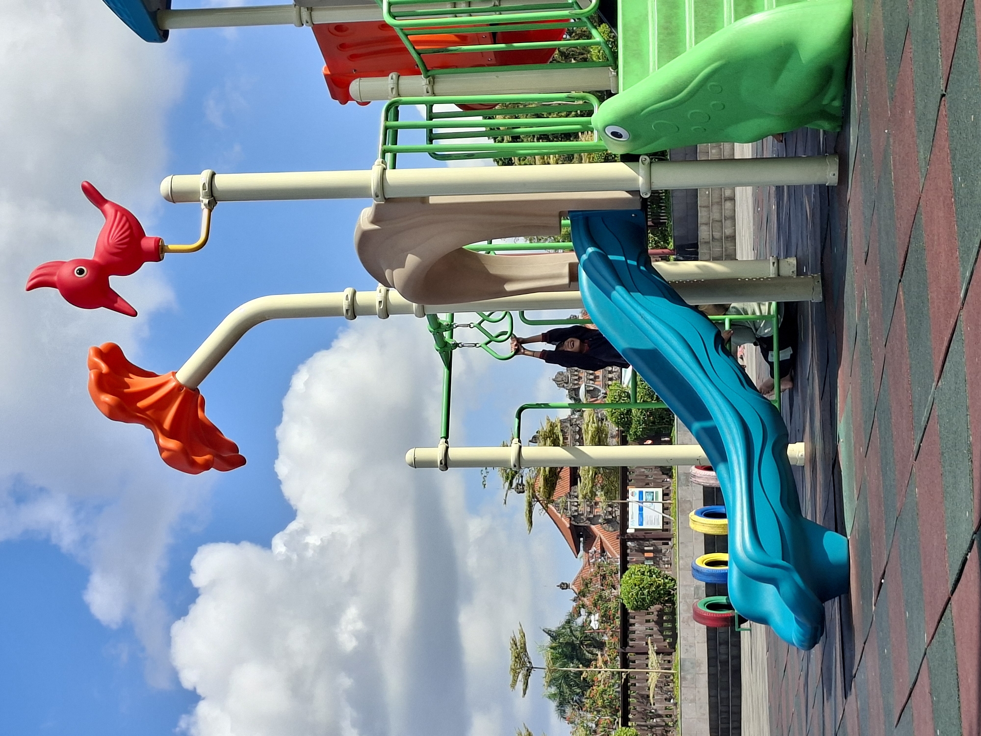 In Gianyar, Indonesia, a brightly colored playground sits under a partly cloudy blue sky. The playground features a blue slide with a frog-shaped base, and a green slide with a frog decoration at its side. In the background, a traditional Balinese temple with intricate architecture and red tile roofs is visible. A child is seen hanging from monkey bars on the playground, their arms extended. The playground is situated on a patterned rubber mat surface.