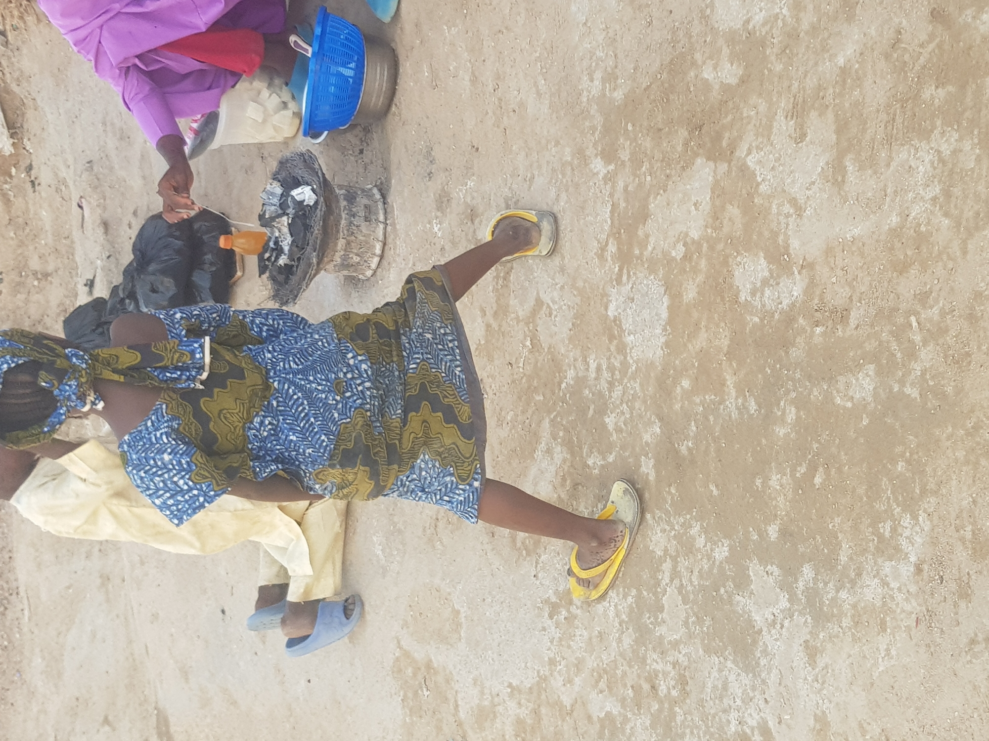 The image depicts two individuals in an outdoor setting in Dabawa, Nigeria. The environment consists of a beige, textured ground. One person wears a blue and green patterned dress and yellow sandals. Another person is wearing a purple top, red pants, and is standing near a charcoal stove. There is a blue basket nearby, as well as a white container, and a black bag next to the stove. One of the individuals seems to be using a thin string to prepare a cooking ingredient above a container of black charcoal and coals. The third person is wearing a cream outfit with blue sandals.