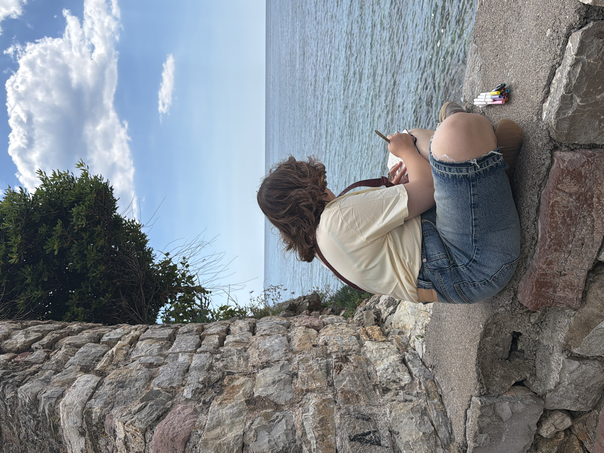 A person is sitting on a stone wall overlooking a body of water, likely the Adriatic Sea, near Budva, Montenegro. The wall appears to be part of a retaining structure or fortification, built with roughly hewn stones. The individual, whose gender isn't immediately clear, has medium-length curly brown hair and is wearing a light yellow short-sleeved shirt, denim shorts with a ripped knee, and brown footwear. They are holding a small notebook or sketchbook in their hands and appear to be drawing or writing with a pen or pencil. Beside them on the wall is a bundle of colorful markers or pens held together by a rubber band. The sky is partly cloudy with blue patches. To the left, vegetation grows on the wall. The focus of the scene is the seated person and their creative activity in a scenic location.