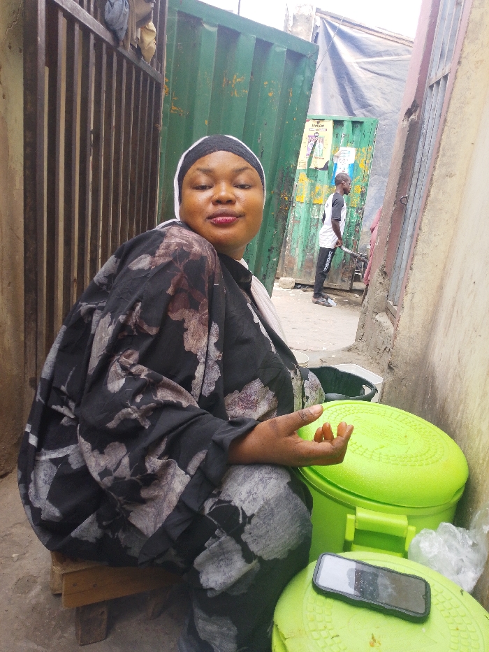 The scene is an outdoor pathway in Fagge A, Nigeria, flanked by various urban structures. An adult female, with light brown skin and a slight smile, occupies the foreground, seated on a low wooden stool. She wears a black floral-patterned loose garment, a white headwrap, and a black outer head covering. Her left hand is raised, revealing reddish-orange staining on the fingertips. To her right are two stacked, bright green plastic containers; a black smartphone rests on the lower lid, alongside a crumpled white plastic bag. In the background, an adult male with dark skin is observed standing, partially facing away from the camera, wearing a white and black shirt and dark trousers, holding a long pole. The immediate surroundings include a vertical slatted metal fence on the left and a beige concrete wall on the right. Further back, two tall, dark green corrugated metal structures, possibly containers or gates, are visible, bearing attached posters. The ground is unpaved.