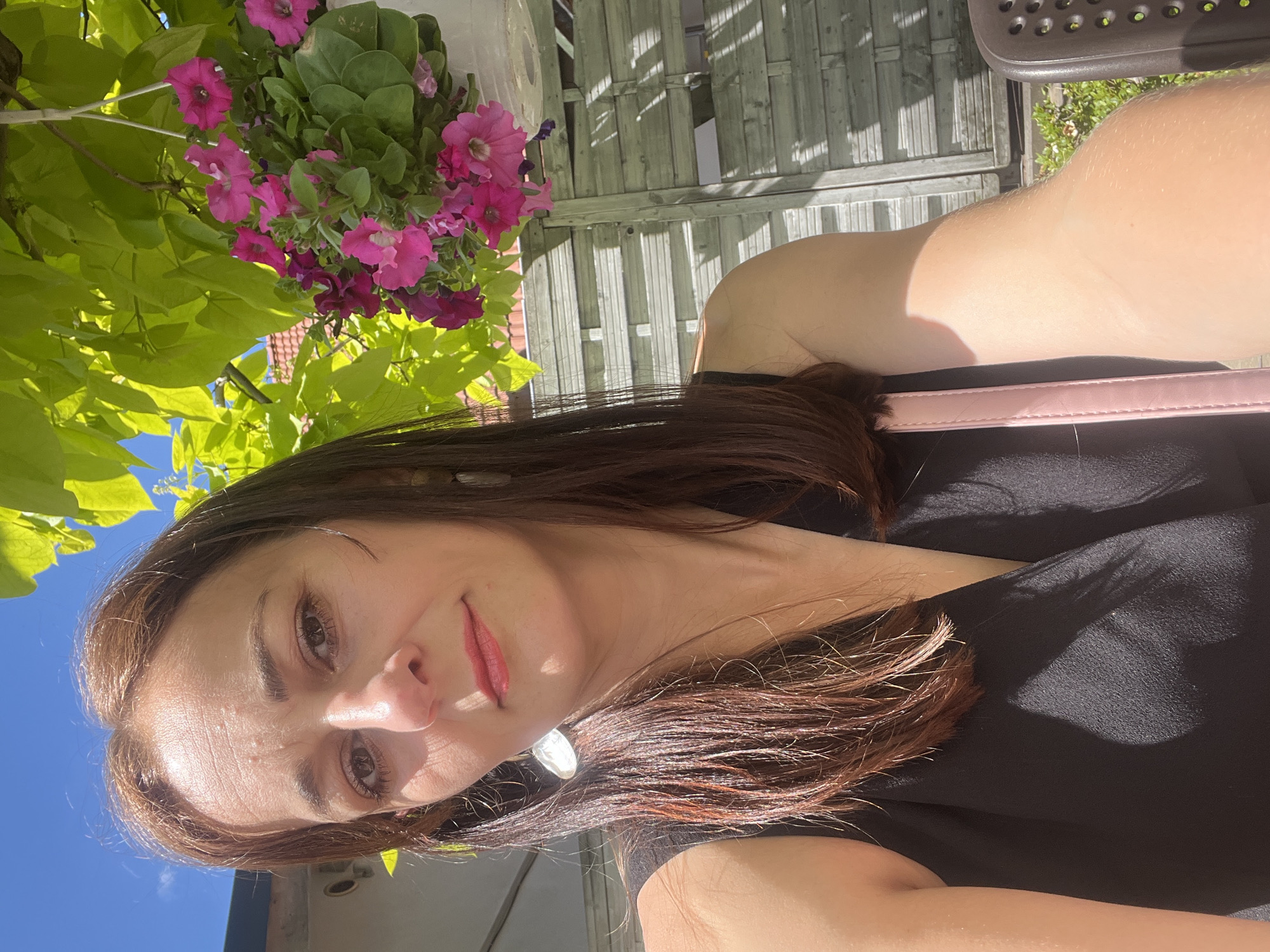A medium shot captured in natural light shows a woman with shoulder-length, dark brown hair and light brown eyes posing for a selfie outdoors. She is wearing a sleeveless black top and a pink strap is visible over her shoulder, presumably from a bag. The woman has red lipstick on and is wearing a large, oval-shaped earring in her left ear. Behind her, a wooden fence with horizontal planks is visible, and above her head, bright green leaves and a hanging basket filled with pink and purple petunias are seen against a clear blue sky. The woman's right arm and shoulder are visible, with sunlight casting a shadow on her arm and the front of her top.