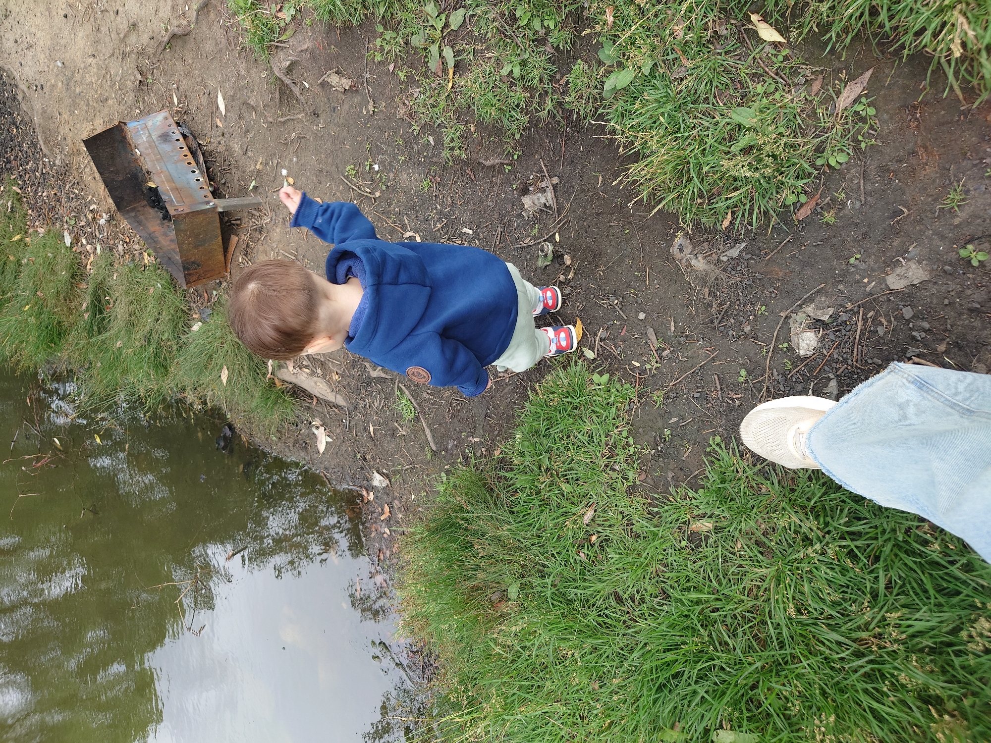 A young child wearing a blue hooded sweatshirt and light-colored pants, with red and white sneakers, stands facing away from the camera near the edge of a body of water. The child is reaching out with their right hand, possibly towards a small white object. To the child's right is a rusty, old metal barbecue grill. The ground is a mixture of dirt, grass, and fallen leaves. In the foreground, a person's foot in a light-colored sneaker and light blue jeans is visible, suggesting the photo was taken by an adult standing near the child. The environment appears to be an outdoor recreational area, likely a park or campsite, situated in the city of Kommunarka, Russia. The water is murky green with reflections of the sky and trees.