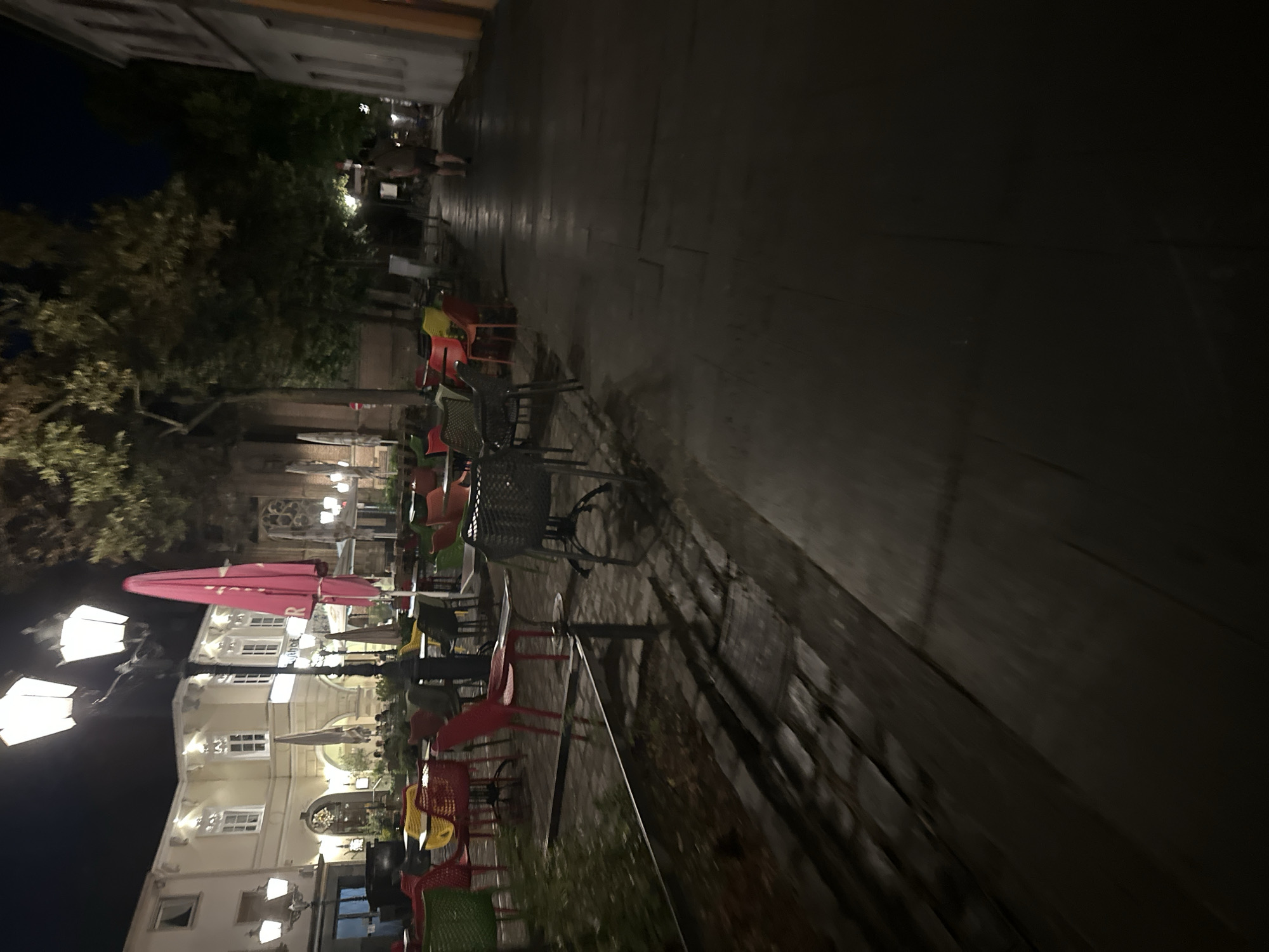 The image depicts an outdoor seating area of a restaurant at night. Several tables are visible, some with chairs, arranged on a cobblestone and paved surface. The seating area is illuminated by lampposts. In the background, a building with multiple windows and architectural details is visible, suggesting it is a historic structure. A person is walking away from the camera on the paved walkway in the middle ground. The overall scene suggests a quiet evening in a European city, possibly Krefeld, Germany, given the context.
