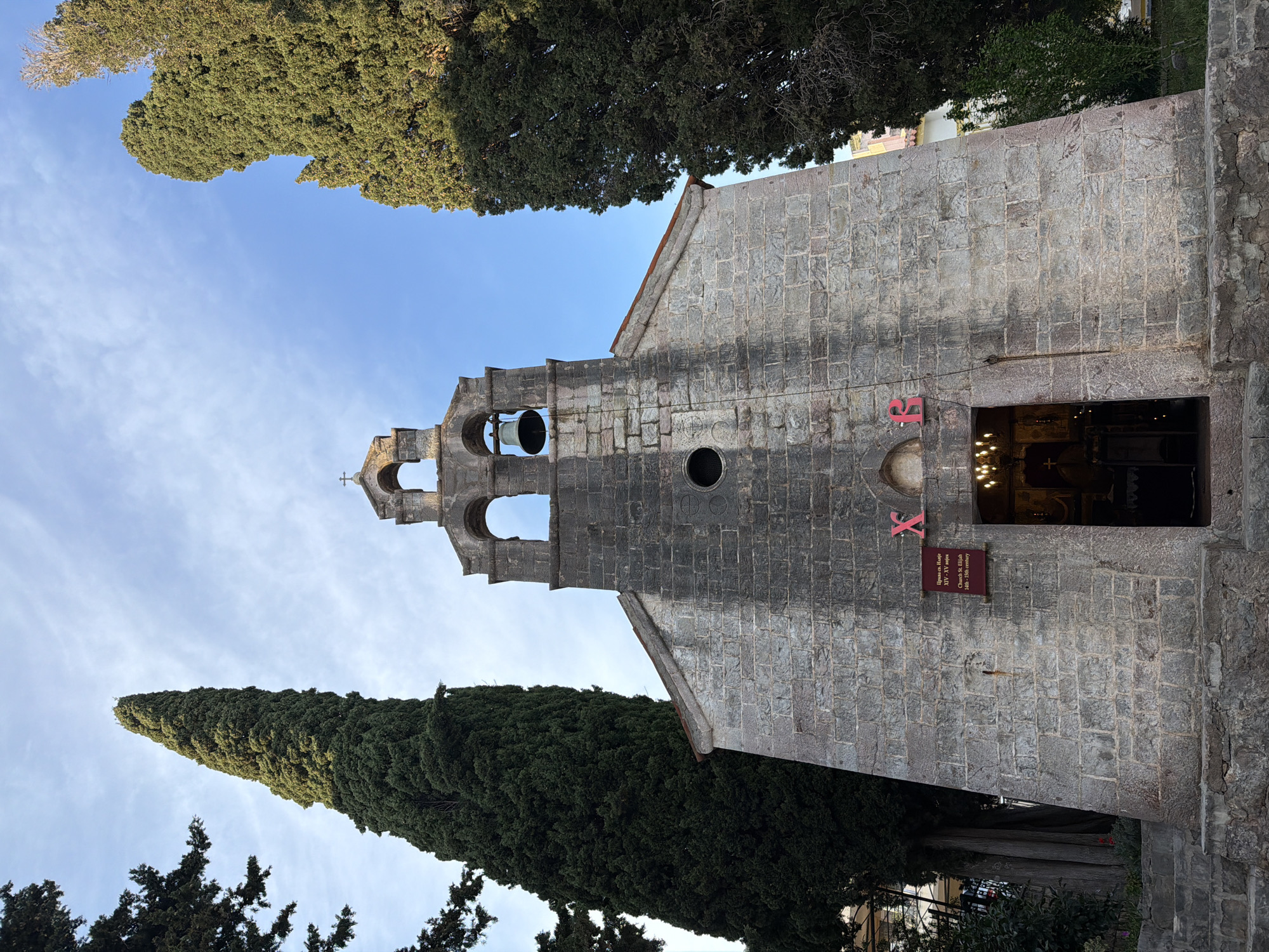 The image shows the Church of St. Elijah in Petrovac centar, Montenegro. The church is built of gray stone and has a bell tower with two bells visible. There's a round opening in the wall next to the tower. Above the doorway, there's a sign in Cyrillic and English that reads: "Church St. Elijah, 14th - 15th century." Two tall, dark green trees flank the church, reaching up towards the blue sky. The picture is taken from below, looking upwards.