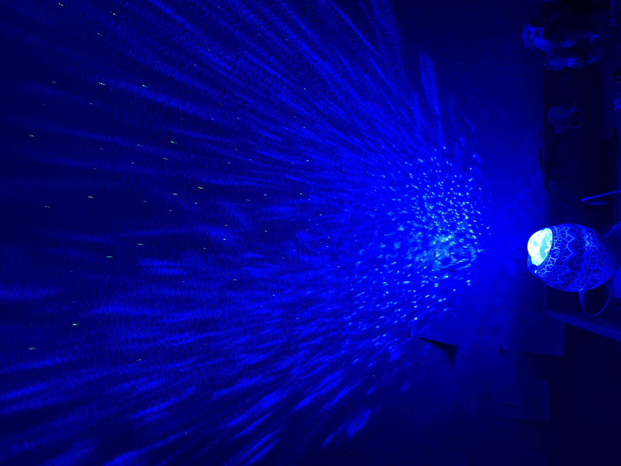 A close-up, eye-level shot shows a blue light projected onto a textured ceiling, creating an ethereal, starry night effect with radial patterns and scattered bright spots. In the foreground, a patterned, egg-shaped lamp with a glowing light inside sits on a dark surface, with wires extending from its base. To the right of the lamp, other objects are dimly visible in the dark environment. The image is devoid of any identifiable landmarks of Colorado Springs.