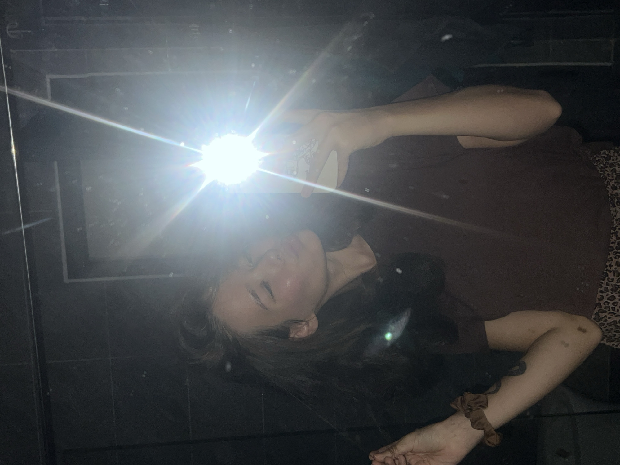 The image displays a person, likely female, taking a selfie while lying down in a low-light setting. The individual is positioned horizontally across the frame, with her head towards the left and her body extending to the right. She has dark hair, and her face is partially illuminated and obscured by a very bright, star-shaped flash emanating from a smartphone held in her left hand. Her eyes appear closed or heavily shadowed by the intense light, making her expression indistinct.
She is wearing a dark brown t-shirt. On her right wrist, a brown scrunchie is visible. Her right arm and lower leg show some faint marks or discolorations. A segment of patterned fabric, possibly leopard print, is visible on her lower right side. The person is lying on a dark, reflective surface, which exhibits faint grid lines suggesting tiles or panels, along with numerous small, bright specks that could be dust or reflections. The surroundings are dark and indistinct, indicating a dimly lit room or nighttime. This scene takes place in Nam Nha Trang Ward, Vietnam. No discernible text is visible in the image.