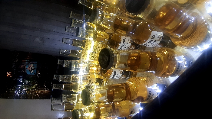 A wall of Corona beer bottles, their amber liquid illuminated by subtle lighting. Most bottles are capped and labeled "Corona Extra." The composition creates an abstract, repetitive pattern. Some light shines through the glass, creating a warm glow. At the left of the image is some vegetation with a blue portrait in the background. A dark angled object intrudes from the lower right.