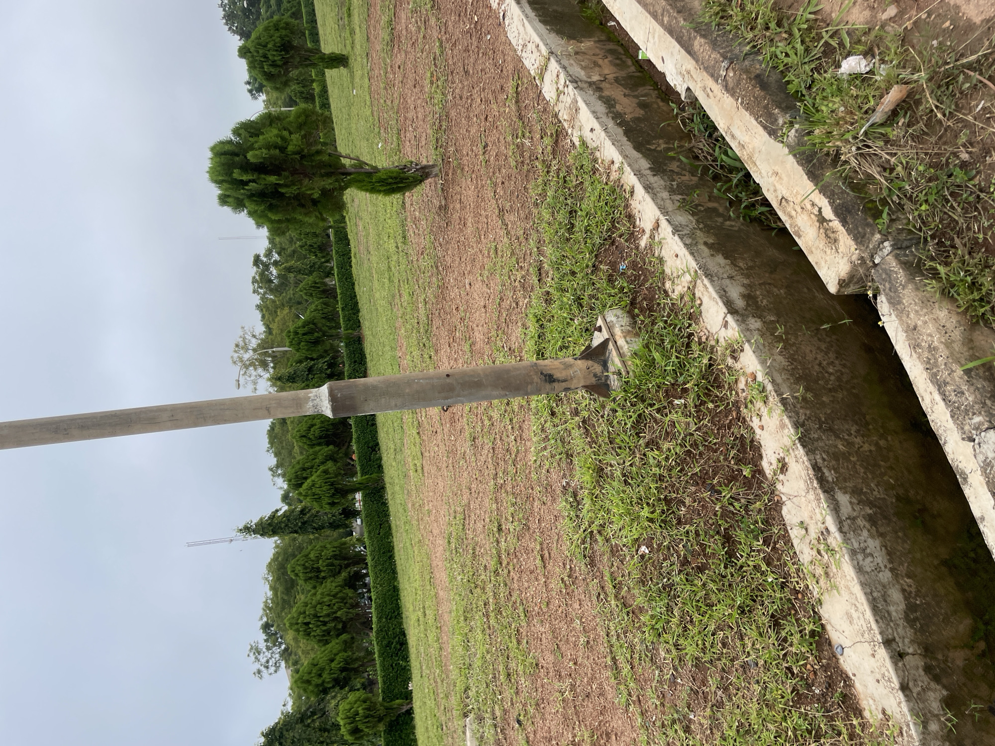 The image depicts an outdoor scene with a concrete drainage channel in the foreground. The channel appears to be partially filled with water and overgrown with vegetation. Beyond the drainage channel, there is a patch of reddish-brown soil with sparse green grass. Further back, a tall, slender pole stands in the middle of a grassy area. In the background, rows of manicured green hedges and various trees are visible, with a cloudy sky overhead. A tall antenna structure can be seen in the distance. The overall environment suggests a park or a large open area within a city, possibly Gwafan, Nigeria, given the context provided. There are no people or animals visible in the image.