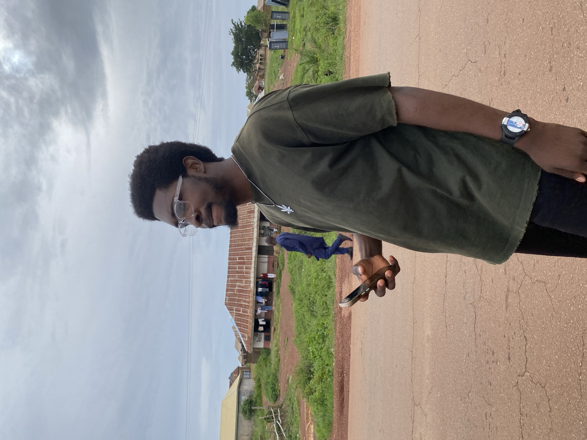 In Durumi, Nigeria, a young Black man wearing glasses and a dark green t-shirt stands on a dirt road. He holds a smartphone in his right hand and a silver necklace with a leaf pendant is visible on his chest. A watch with a black strap is on his left wrist. In the background, several buildings with corrugated metal roofs are situated amidst lush green vegetation. A man in a blue outfit walks away from the camera in the middle ground, and clothes are hanging to dry outside one of the buildings. The sky is overcast with a cloudy sky.