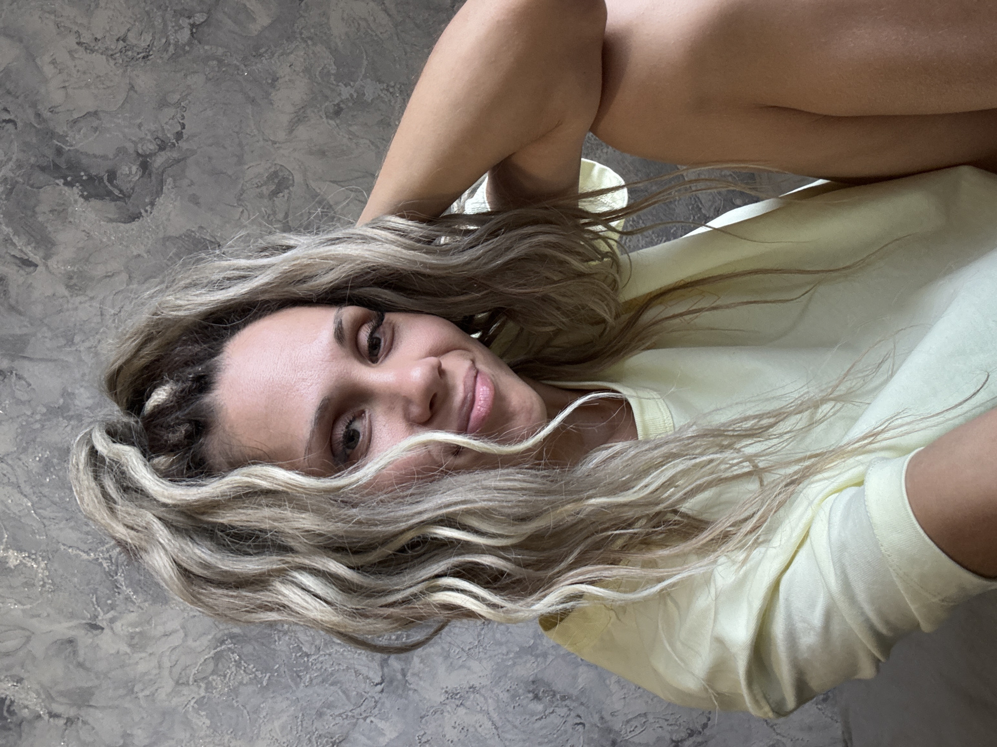 A woman is pictured in a close-up portrait shot, positioned on the right side of the frame. She has blonde, wavy hair that is partially covering her face. She is wearing a light yellow t-shirt with the sleeves rolled up. Her right arm is bent and her hand is resting on her head. The background is a textured grey wall. The image does not contain any information about the city of Rusanovo, Russia, or any specific location details.