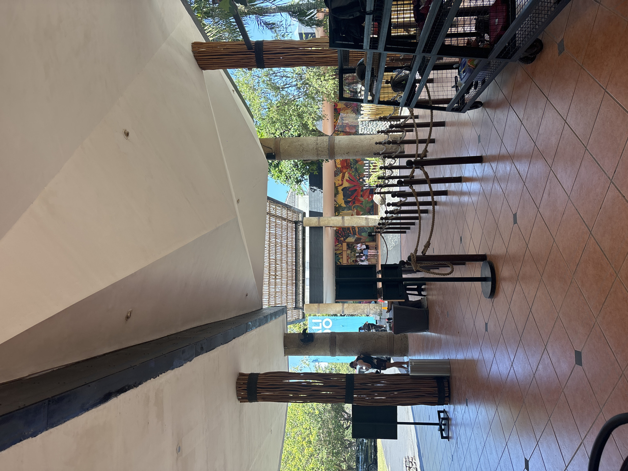 The image shows an outdoor covered walkway at the Bali Zoo in Sukawati, Indonesia. The walkway has a tiled floor and a large, sloped roof supported by columns. Some of the columns are wrapped with natural wood. There are ropes and stanchions forming a queue line, and a metal cart filled with bags is visible on the right. In the background, there is a colorful mural depicting animals. Several people are present, including a person leaning against a column and a small group of people standing together. The environment appears bright and sunny, with trees and a blue sky visible beyond the walkway.