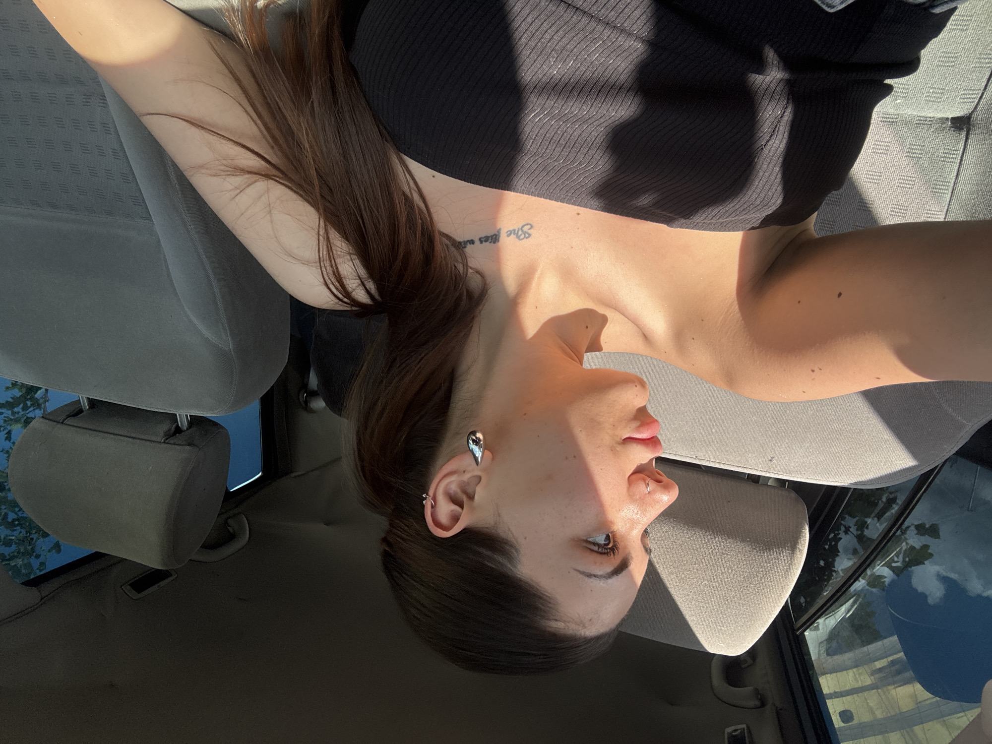 A young adult female is depicted inside a vehicle, positioned in the passenger seat and looking out the left side window. She has dark brown hair styled in a low ponytail and is wearing a black strapless top. Her left ear features multiple piercings, including a large, silver-colored teardrop-shaped earring in the lobe. A small silver hoop nose ring is visible on her left nostril. A tattoo displaying the partial text "She flies with" is present on her right collarbone. Strong sunlight illuminates her left side, casting shadows across her face and upper torso.
The car's interior features light grey fabric upholstery with a textured pattern on the seats and headrests. The roof liner is dark-toned and appears worn, with a grab handle visible above the rear passenger door. Through the rear and side windows, a clear blue sky with some white clouds and green tree foliage is visible, with reflections of what appears to be a building structure in the rear window. The scene is situated within a vehicle in the city of Sofia, Bulgaria.